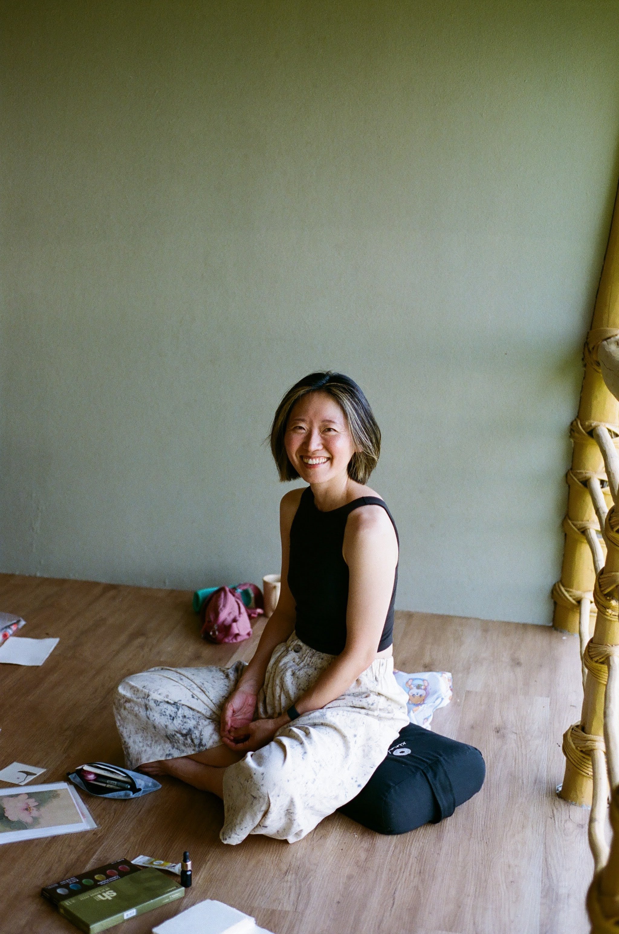Two women sitting on the floor, smiling, with makeup products and papers around them, in a room with green walls and wooden flooring.
