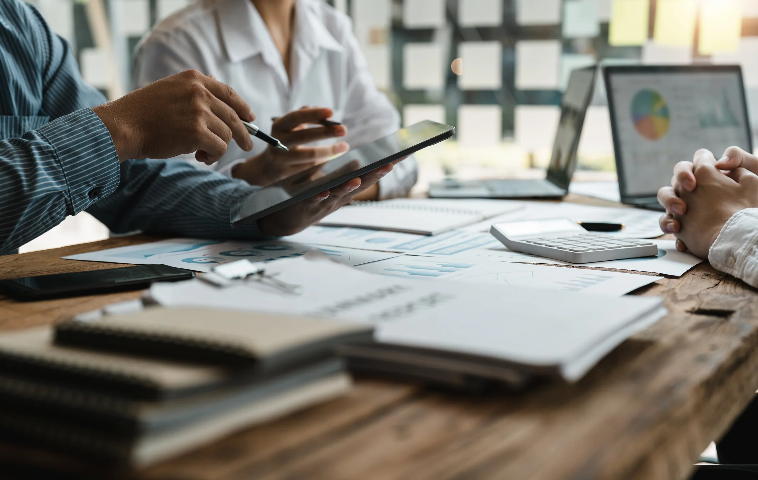 People in business attire collaborating at a meeting, with documents, a calculator, and electronic devices on a wooden table.