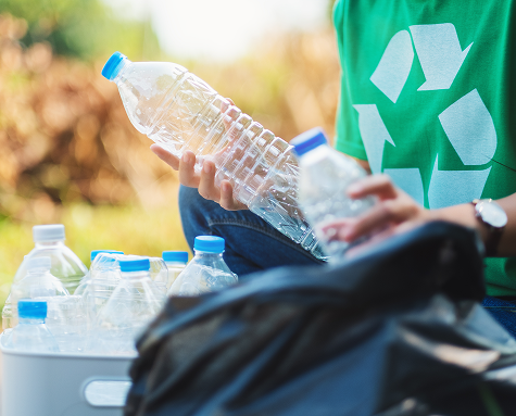 Person placing a plastic bottle into a recycling bin, wearing a green shirt with a recycling symbol, outdoors with other plastic bottles nearby.