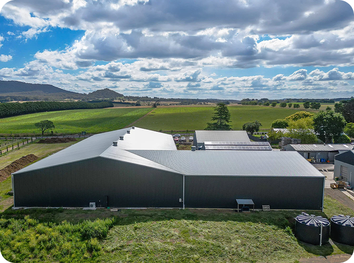 A large industrial barn with a metal roof situated in a rural farmland landscape under a partly cloudy sky.