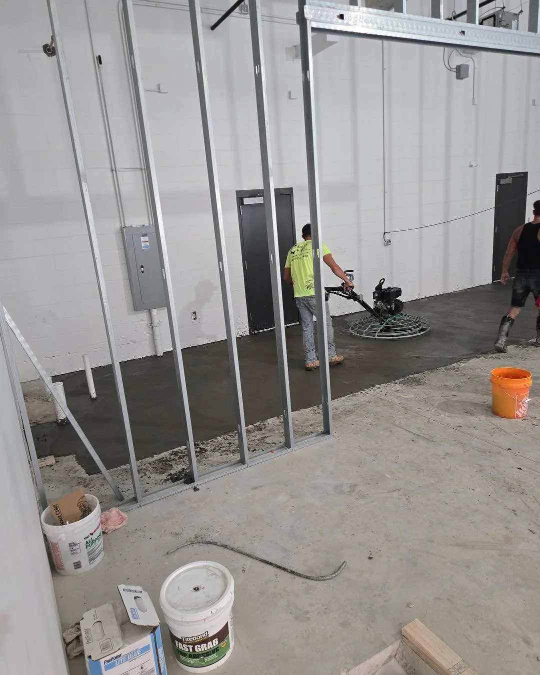 Construction workers pouring concrete inside a building with metal framing. One worker uses a power screed, and another walks nearby. Construction materials and tools are visible, including buckets, cement, and a wooden plank.