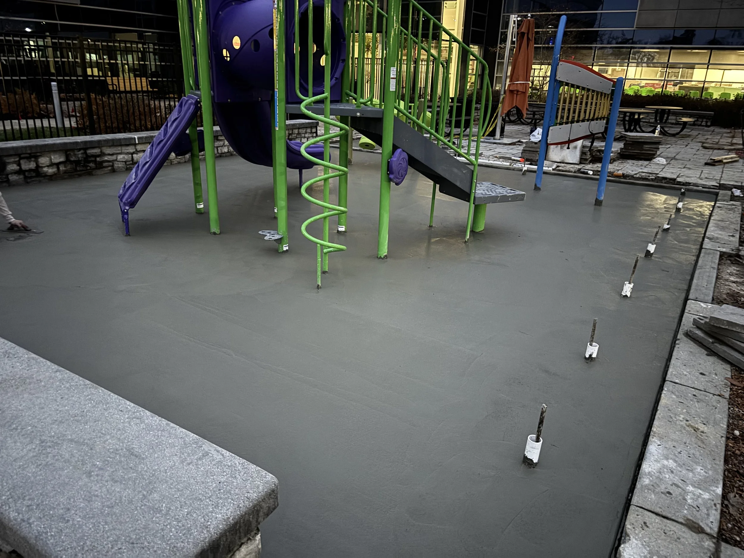 A playground under construction with freshly poured concrete surface, partially assembled colorful play equipment, and construction tools and materials nearby.