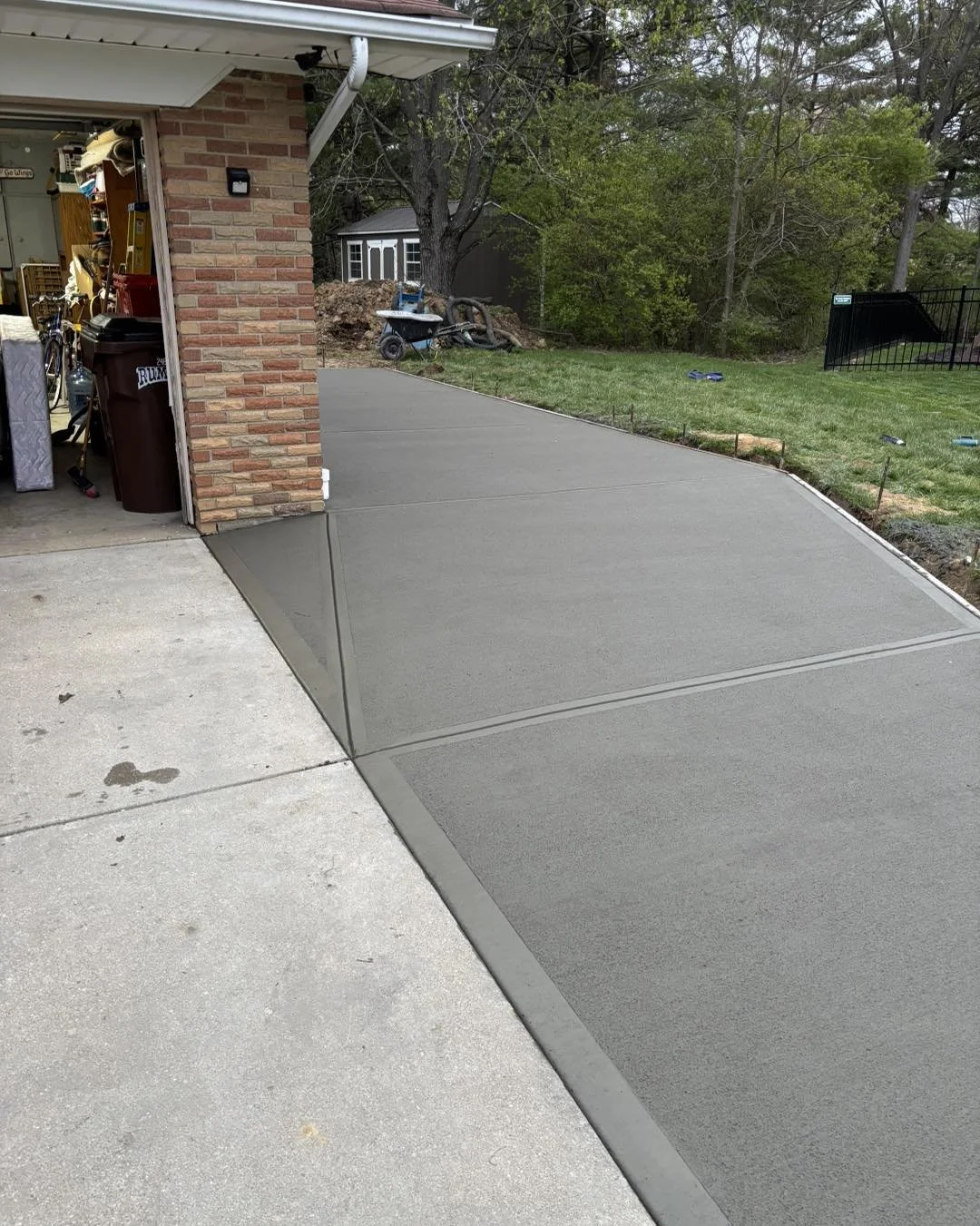 Freshly poured concrete driveway expanding from garage to yard, with visible curb and smooth surface, near a brick house with open garage door.