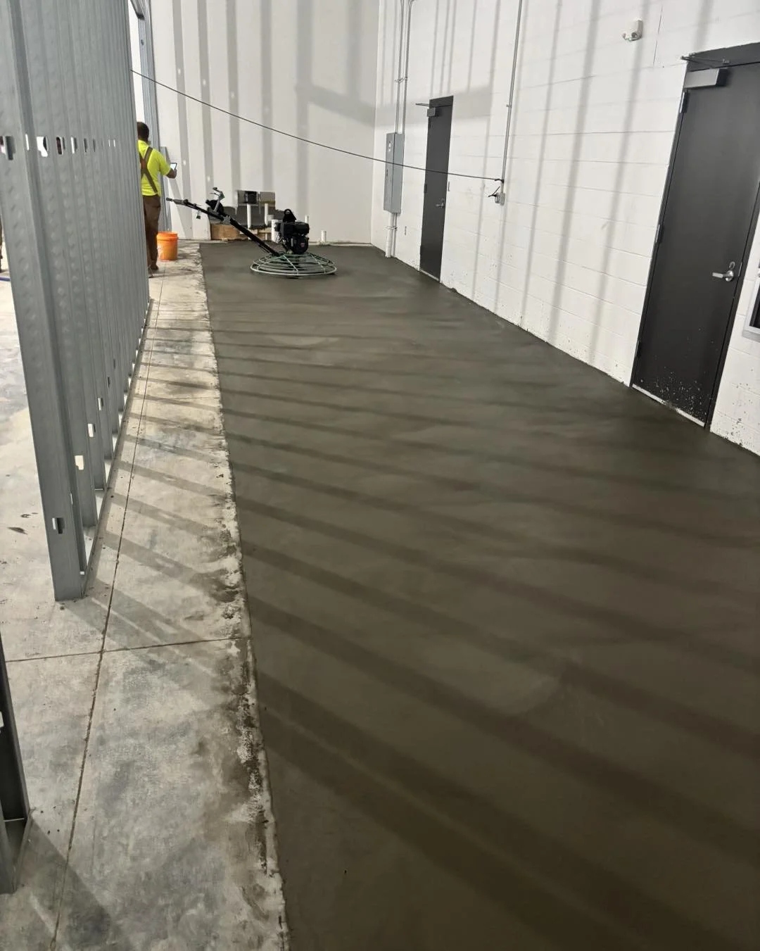 Indoor construction site with a worker using a power trowel on wet concrete floor, surrounded by construction materials and tools.