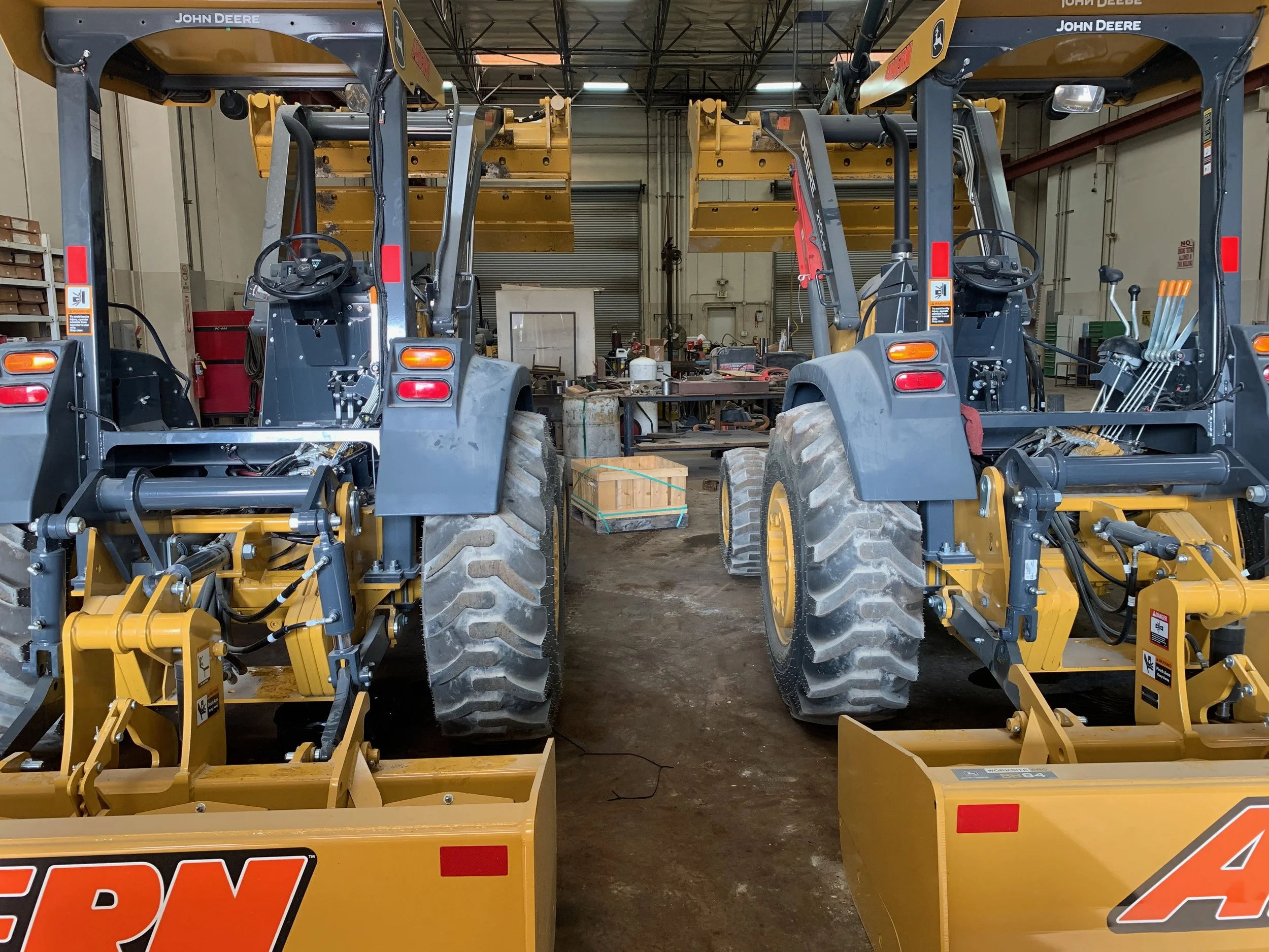 Two yellow John Deere tractors with front blades, parked side by side inside a workshop, with tools and equipment in the background.