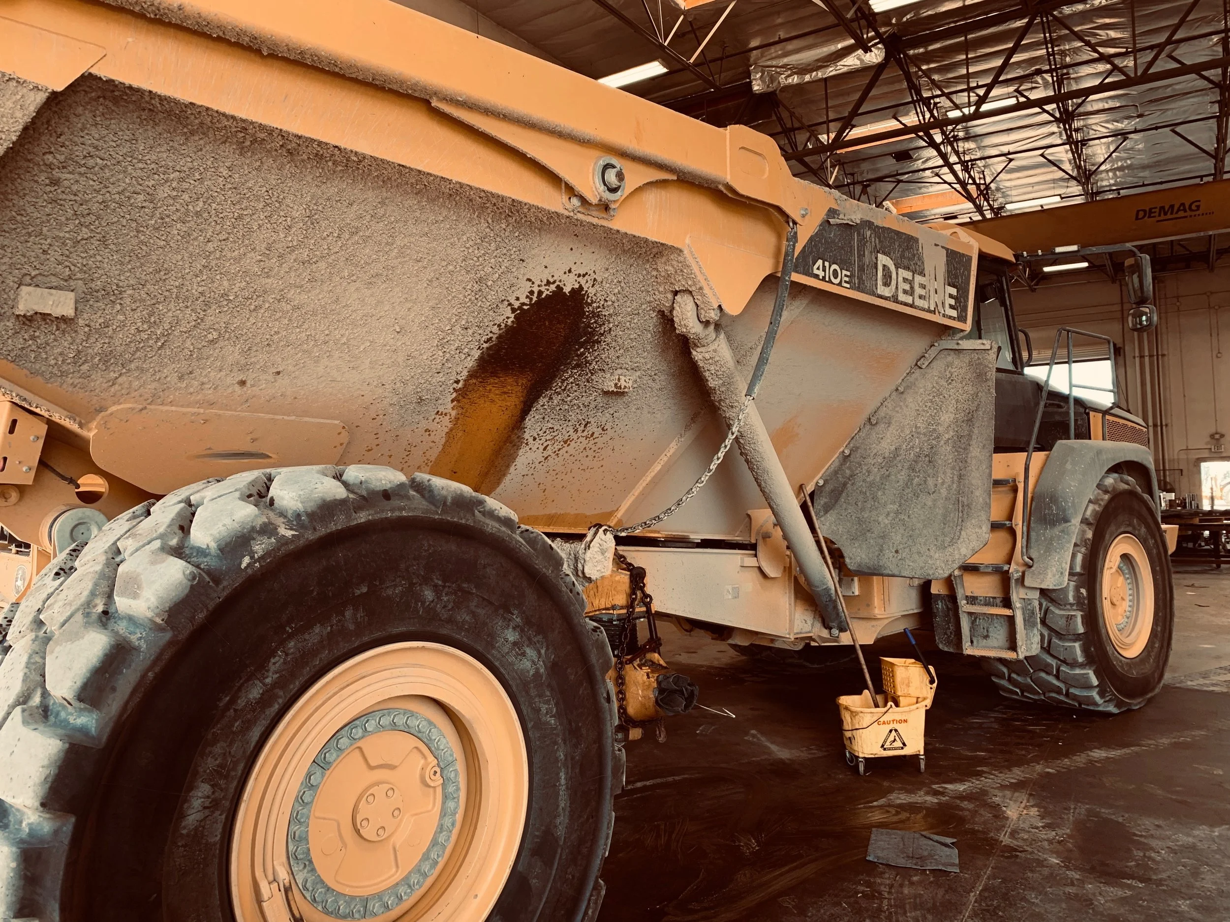 A large construction vehicle, specifically a John Deere 410E dump truck, parked inside a maintenance facility with a black and orange color scheme, dirty tires, and catalytic containers nearby.