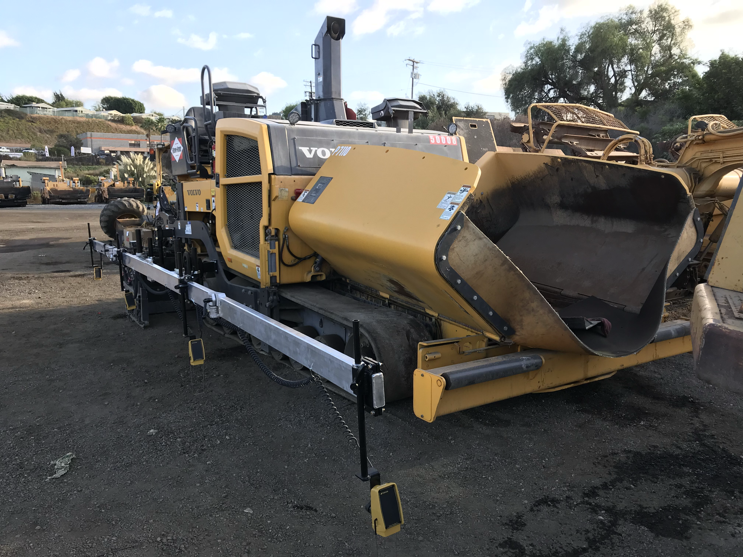 A large yellow and black road paving machine on a construction site, with dirt and other construction equipment visible in the background.