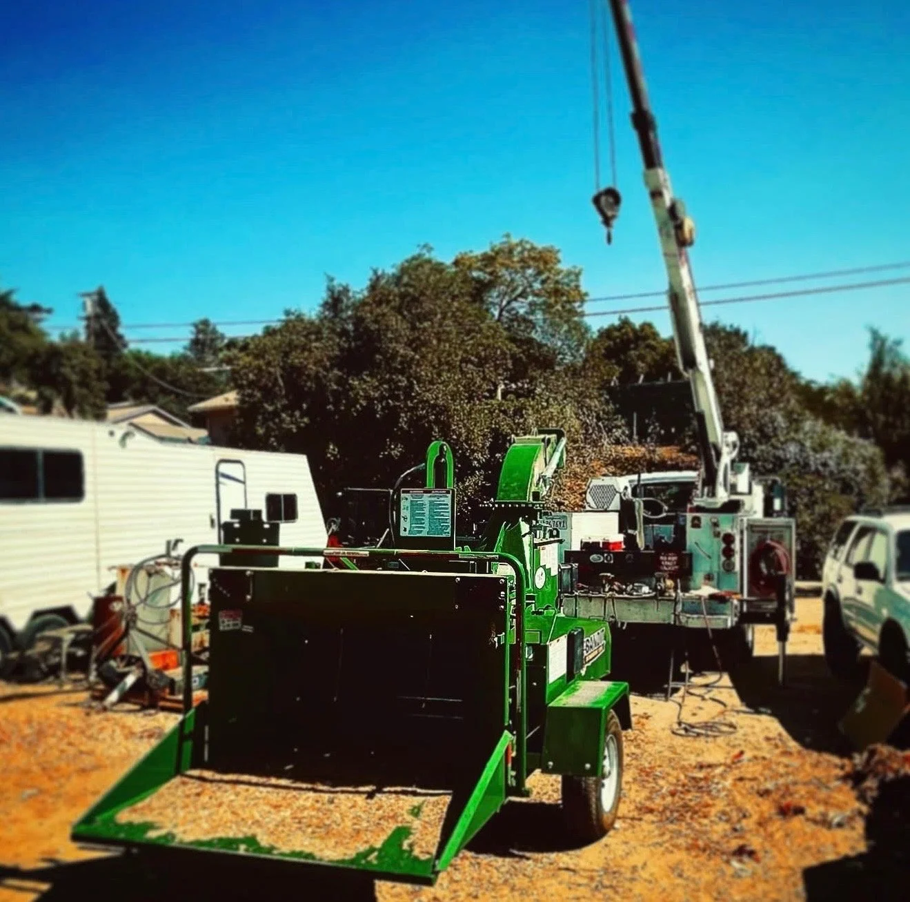 A green wood chipper parked outdoors on a dirt surface, with a white truck crane and vehicles in the background under a blue sky.