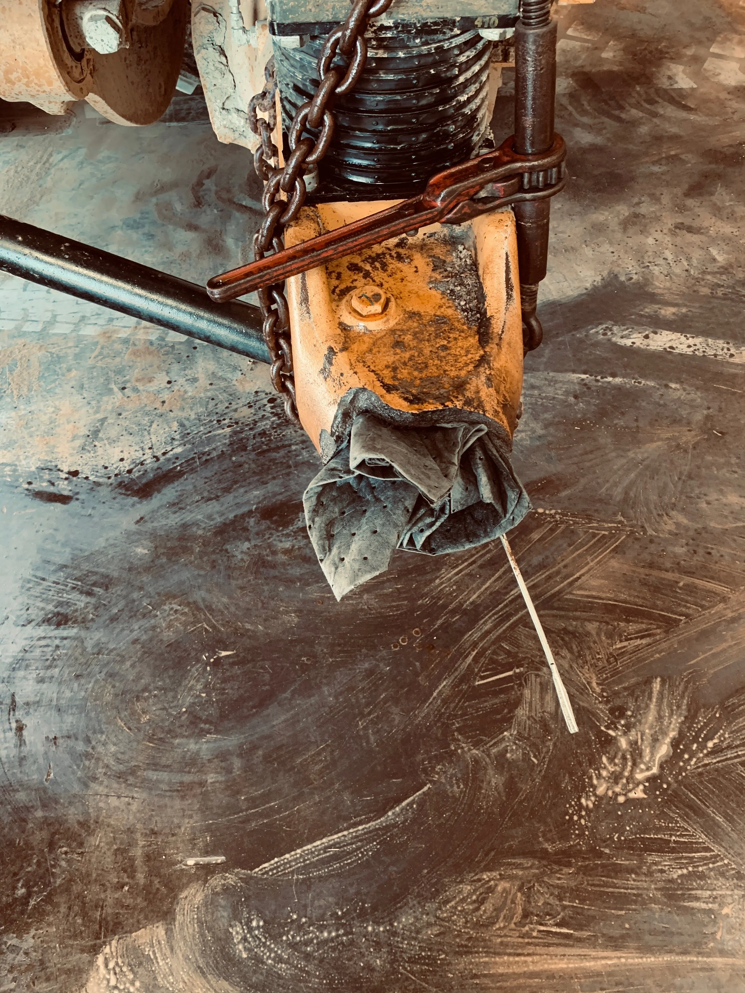Close-up of a rusty metal assembly with chains, a black coil spring, and a dirty, worn orange-painted metal part. The background surface is a dark, textured floor with visible scuffs and marks.