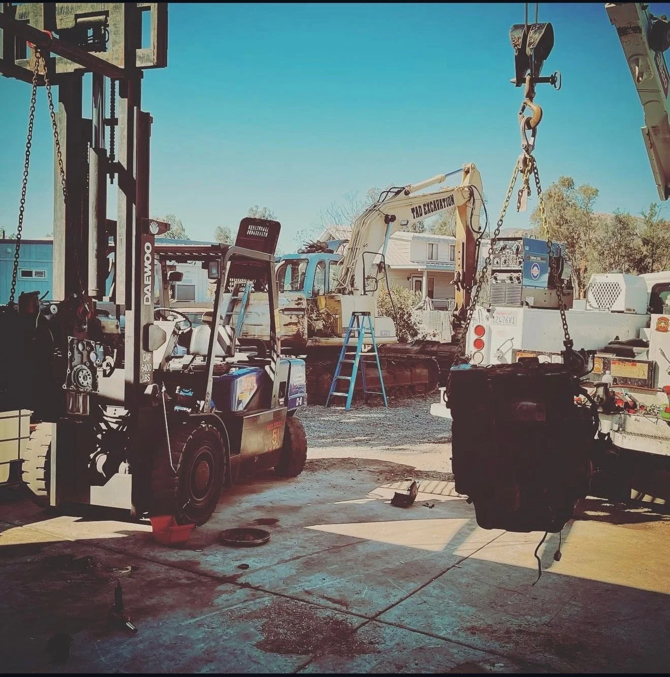 Construction yard with machinery including a forklift, a small excavator, and a crane with chains hanging, under a clear blue sky.