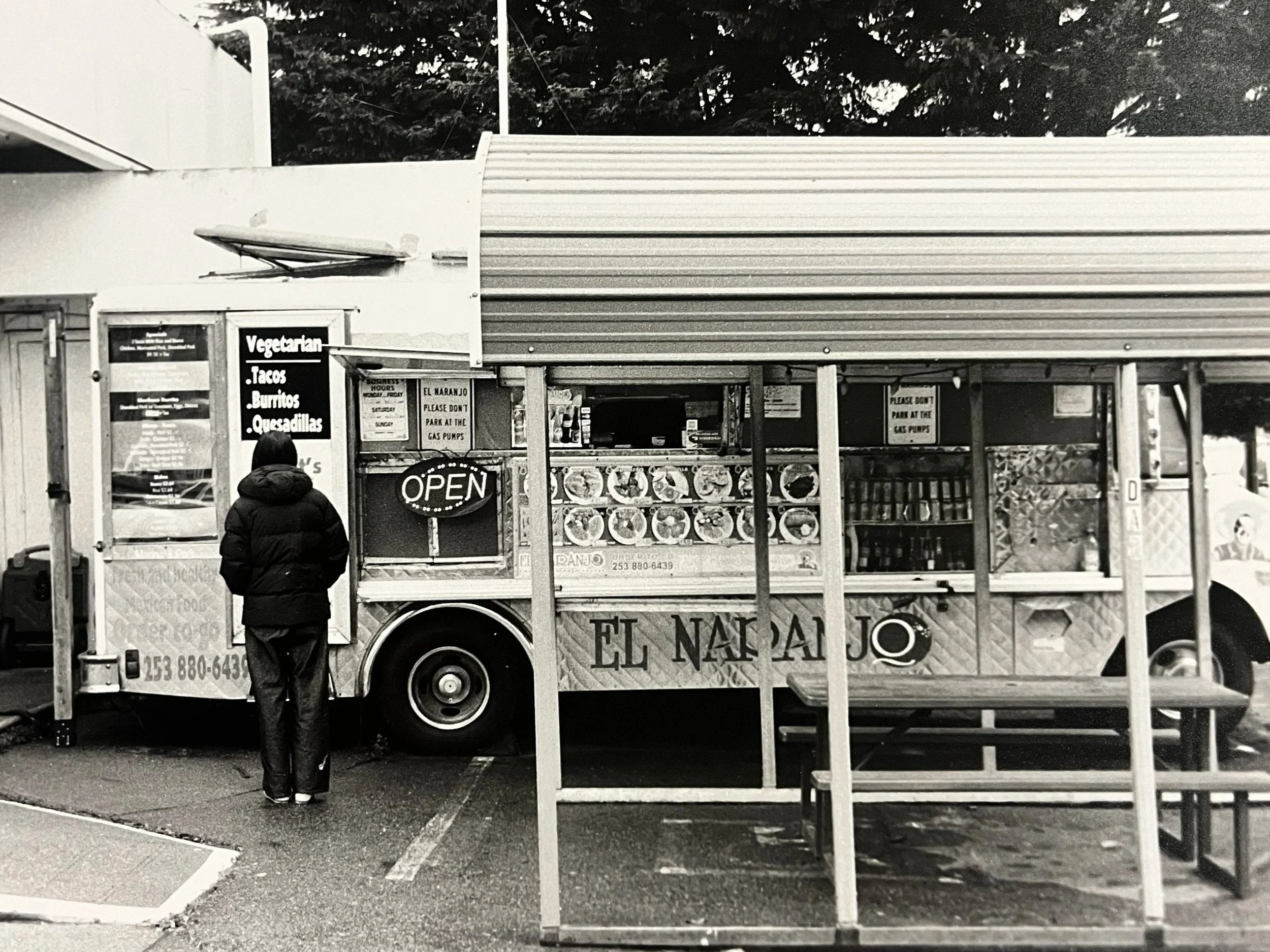 A black and white photo of a food truck with a person standing in front of it. The food truck displays a menu with vegetarian options including tacos, burritos, and quesadillas. An illuminated sign indicates the truck is open, and various packaged fo