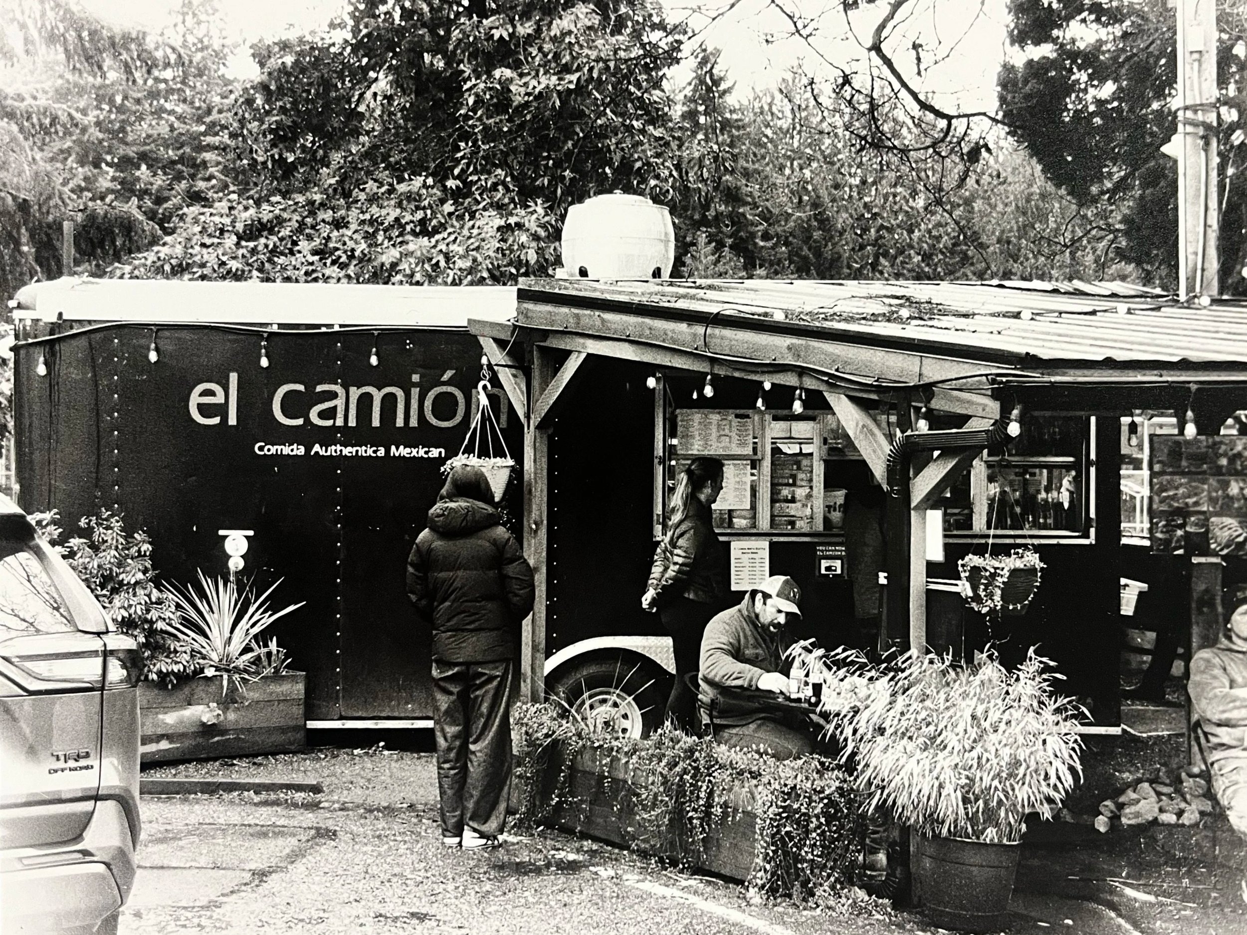 Black and white photo of a Mexican food truck named 'el camión' with a sign that reads 'Comida Auténtica Mexicana'. Several people are in front, one person ordering, and one sitting at a small table. The truck has string lights and potted plants arou