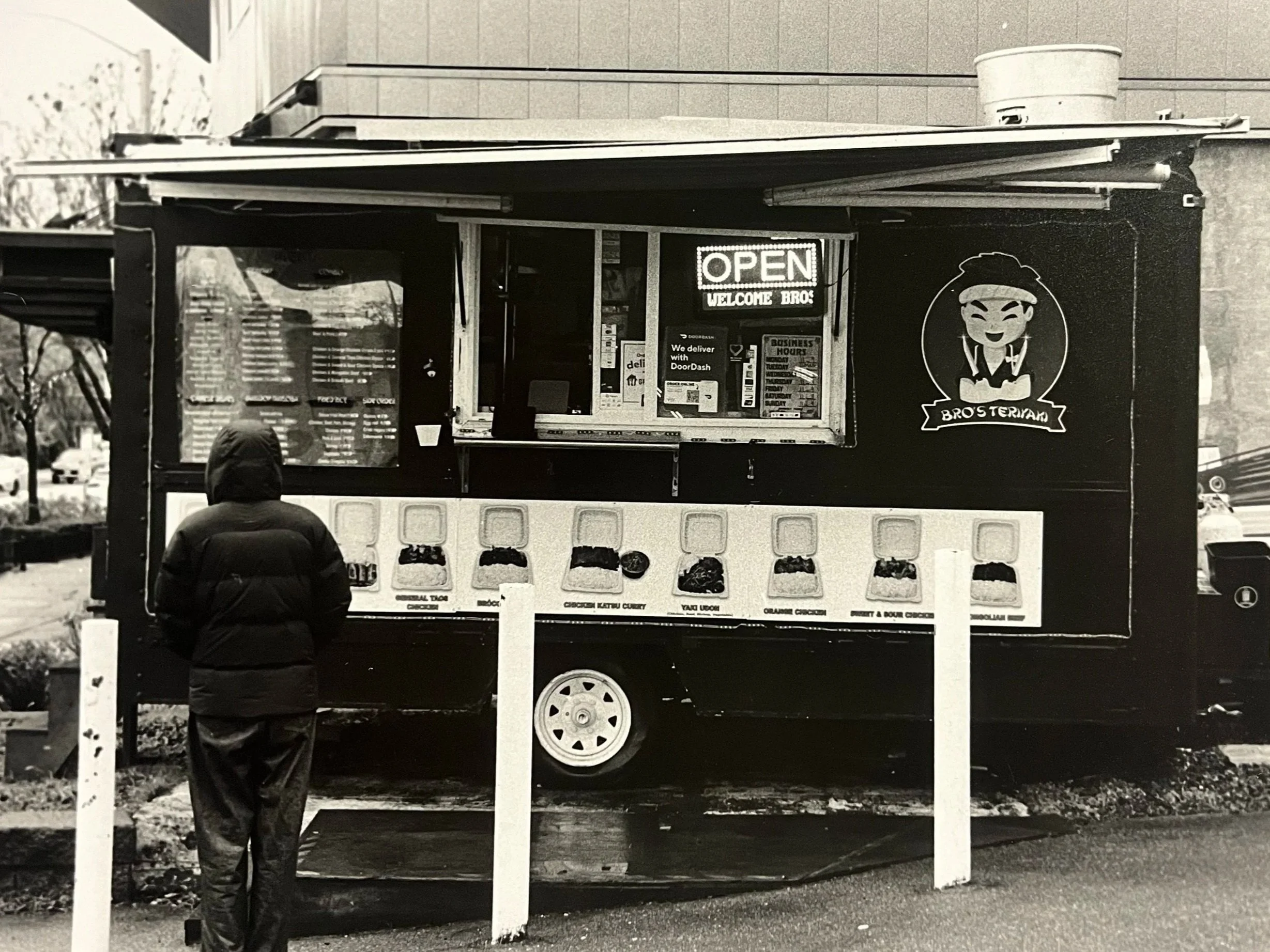 A person in a hooded jacket standing in front of a food truck with a sign saying 'OPEN' and 'WELCOME BRO'.