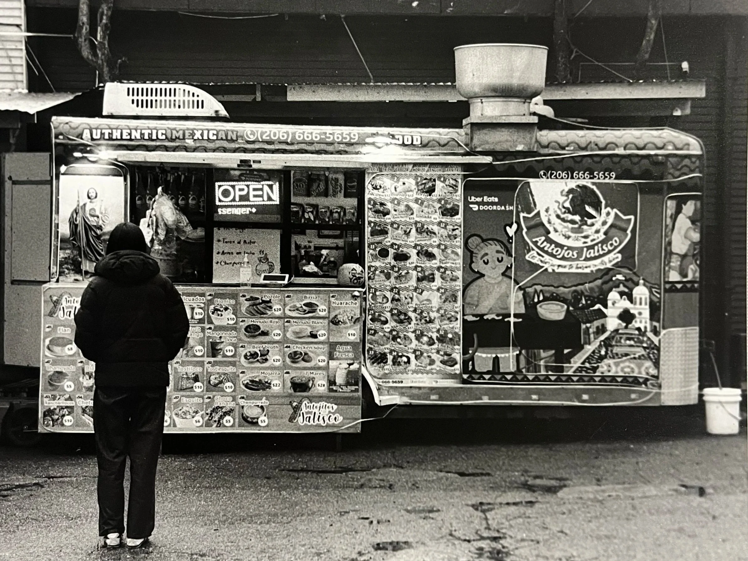 A person standing in front of a Mexican food stand with colorful menu, sign that says 'OPEN', and decorative food imagery on the side.
