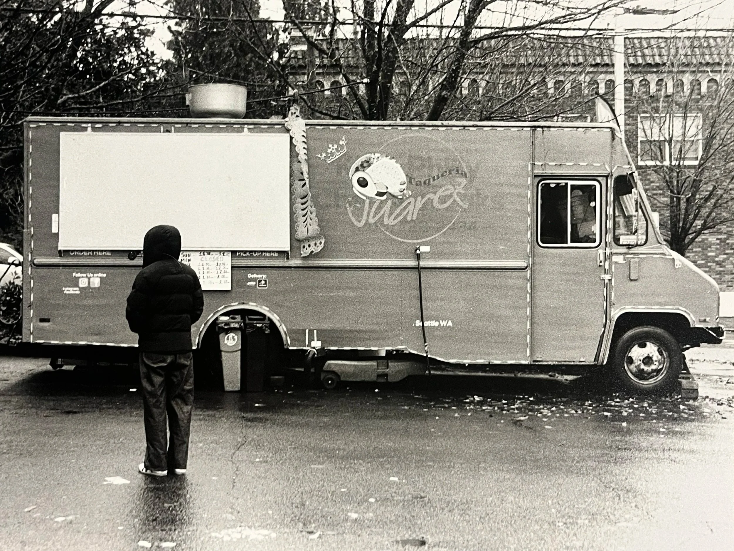 A person wearing a hooded jacket standing in front of a food truck with a colorful logo, on a wet street with fallen leaves and bare trees in the background.