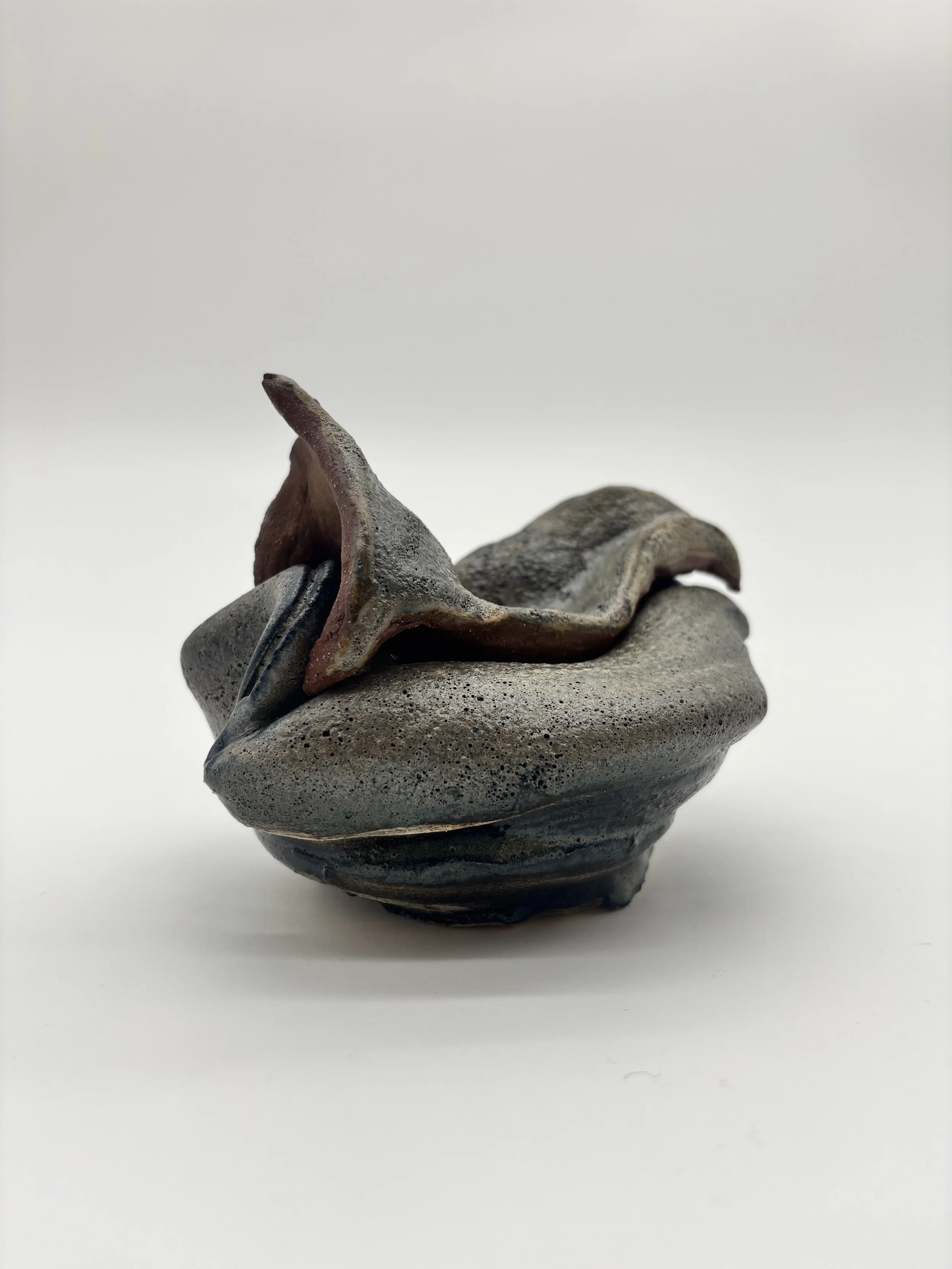 A small, abstract ceramic sculpture with a smooth, layered, rock-like appearance and a curved protruding element on top, set against a plain white background.