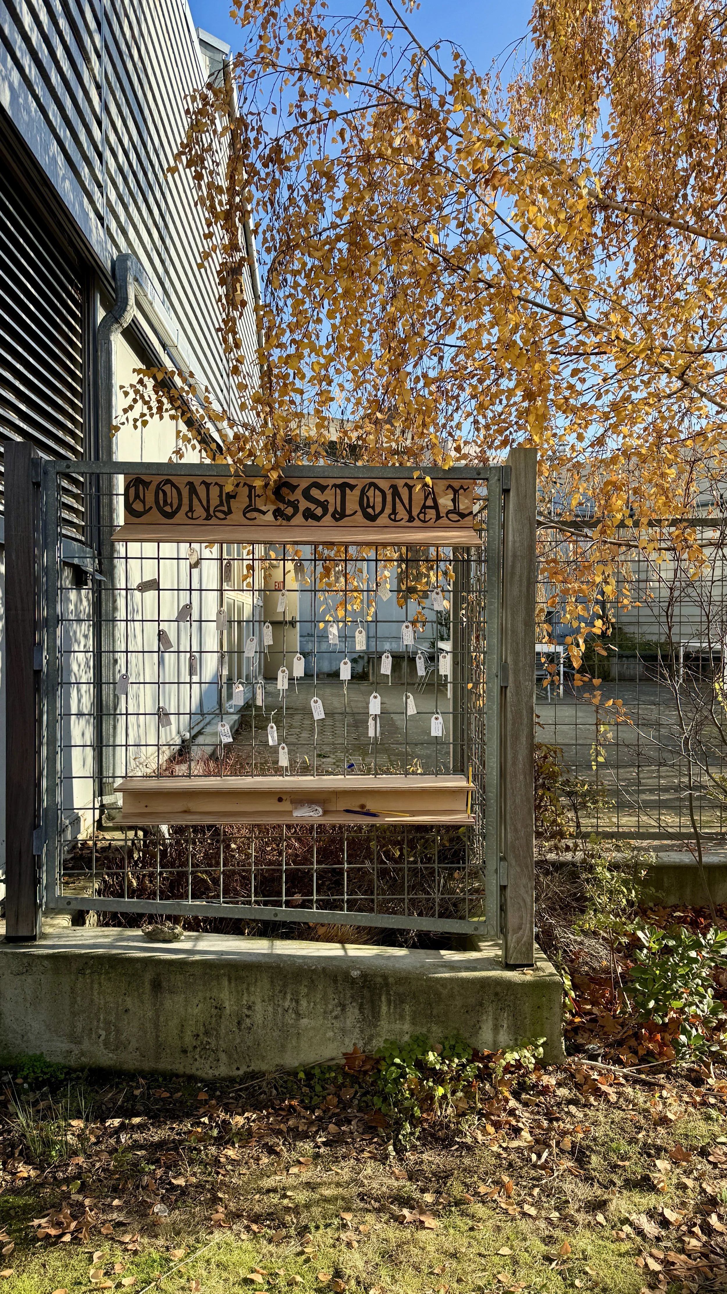 Outdoor gate with a wooden sign that says 'Confessional' and small tags hanging from strings behind the gate. Fall foliage in the background.