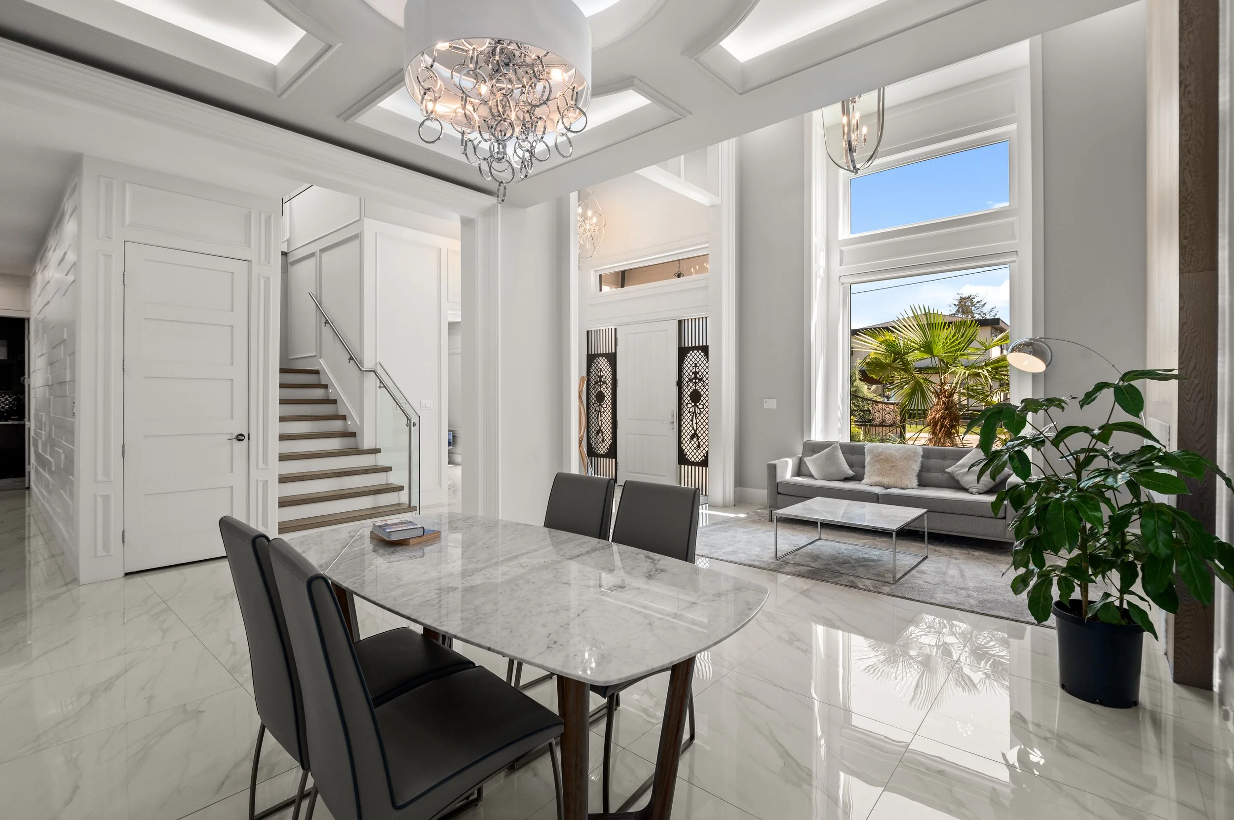 Bright, modern living room with white walls, high ceilings, large windows, a gray sofa with white pillows, a marble dining table with four black chairs, a large potted plant, and a staircase with wooden steps and glass railing.