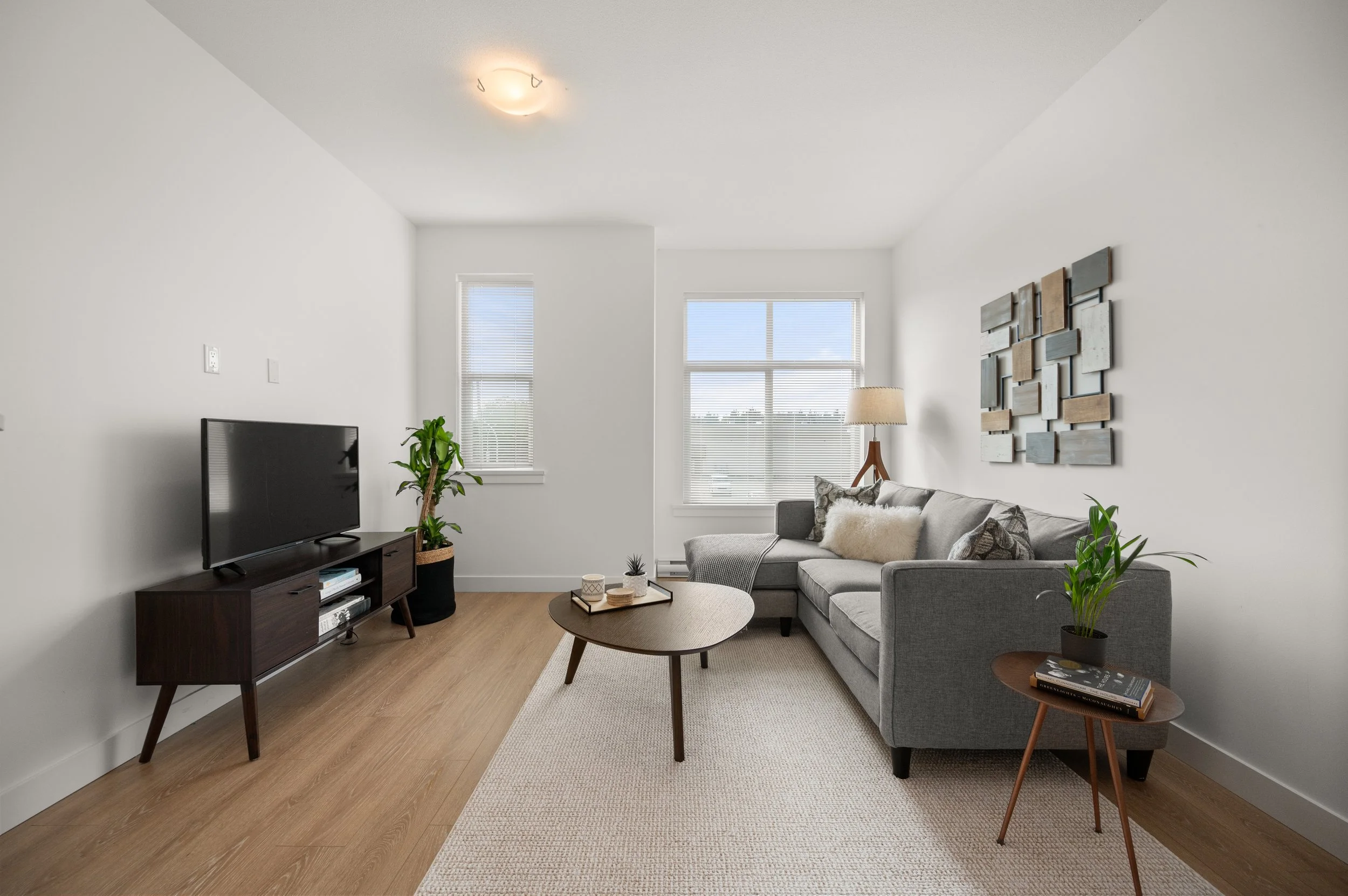 Modern living room with gray sectional sofa, wooden coffee table, flat-screen TV on a wooden stand, potted plants, side table with magazines, framed wall art, large windows with blinds, and carpet over hardwood floors.