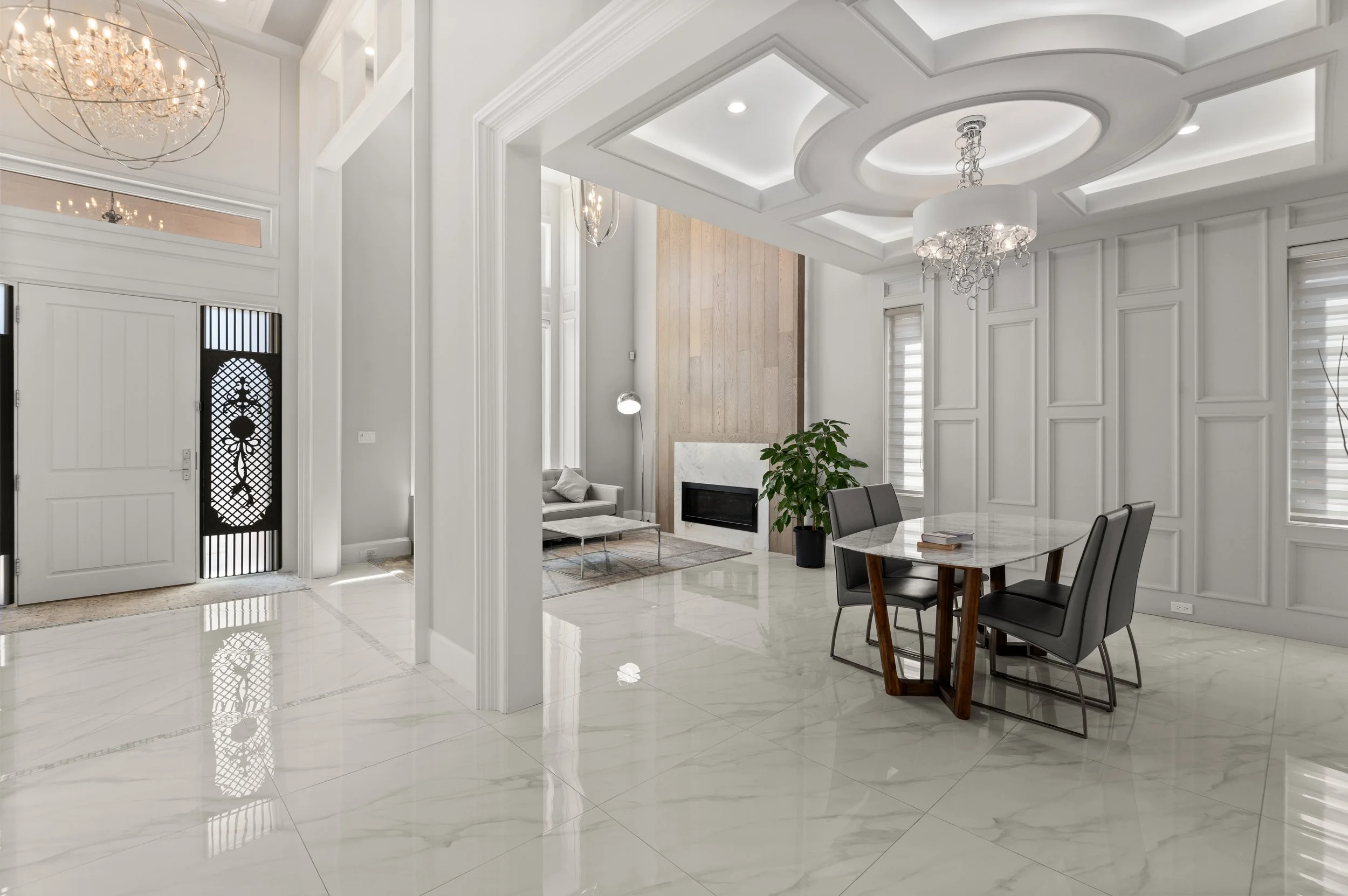 Modern interior with marble floors, white panel walls, a round dining table with four black chairs, and a sitting area with a fireplace and a sofa.