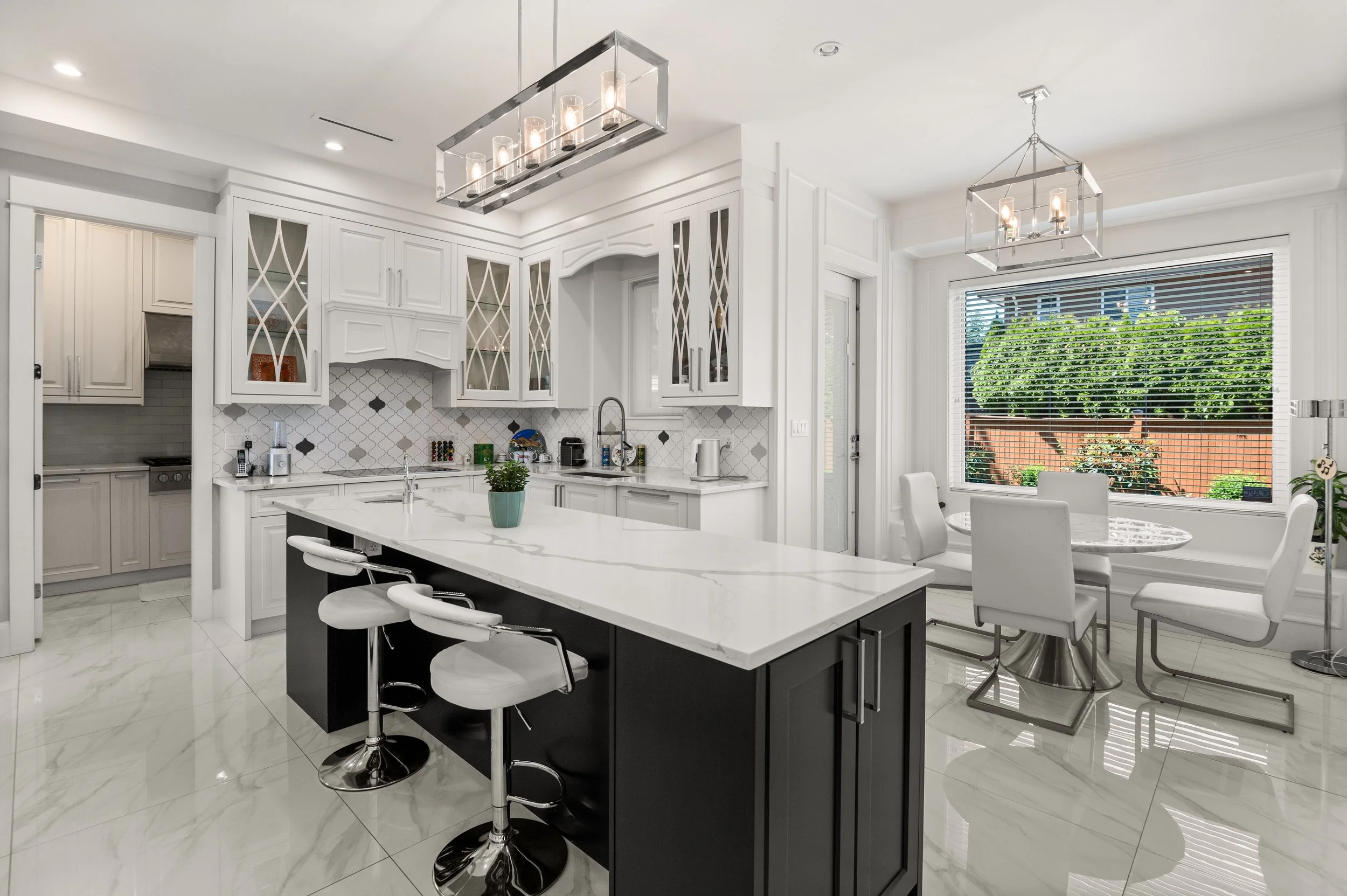Modern white kitchen with black island, marble flooring, glass cabinets, and a dining area with white chairs and a window view