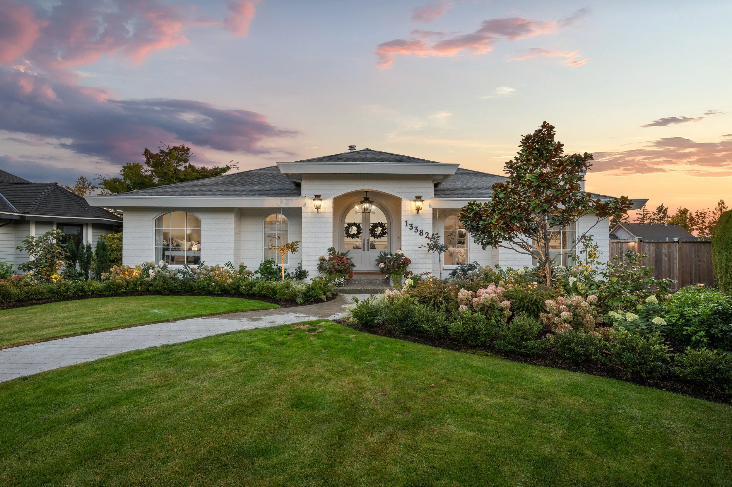 White house with arched front door decorated with Christmas wreaths, front yard with lush green grass, flower beds, small trees, and sunset sky.