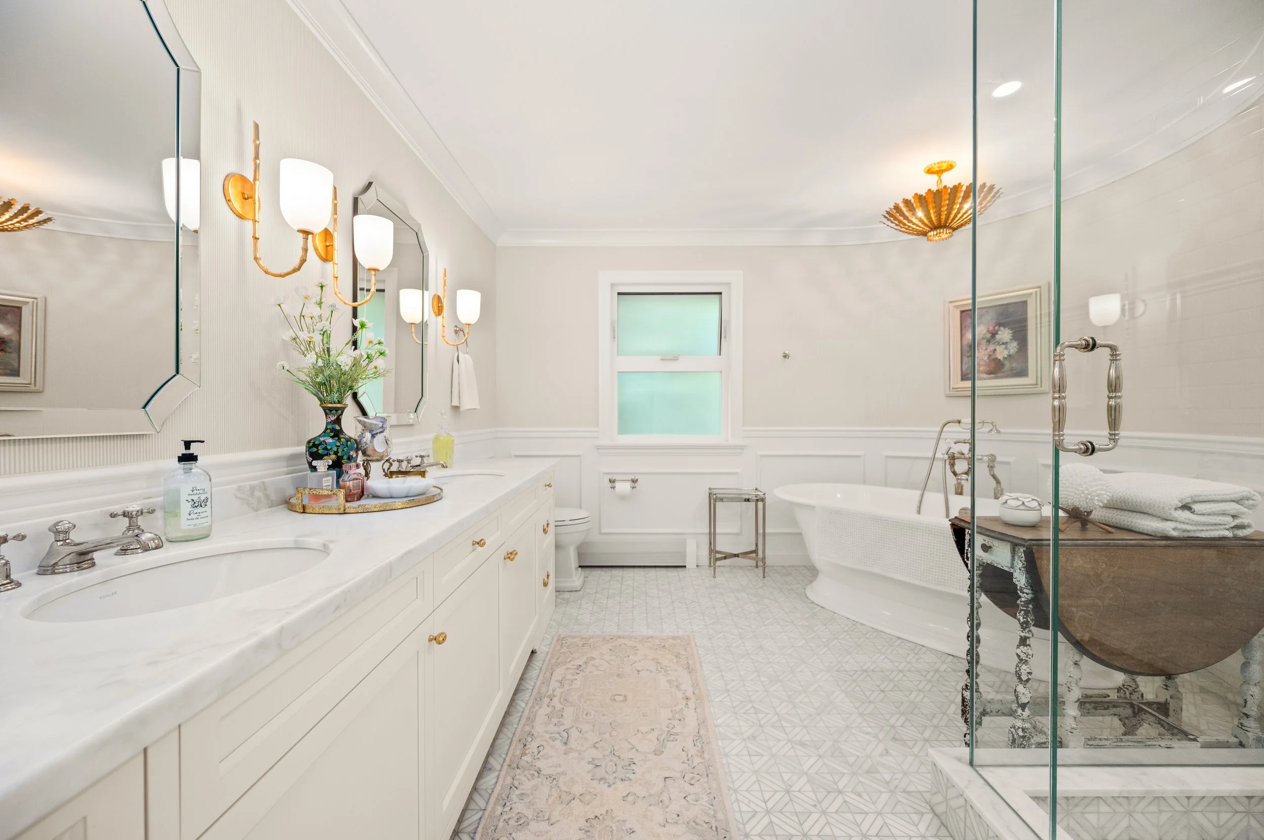 Bright, elegant bathroom with double vanity, large mirror, gold fixtures, flower vase, bathtub, and glass shower enclosure.