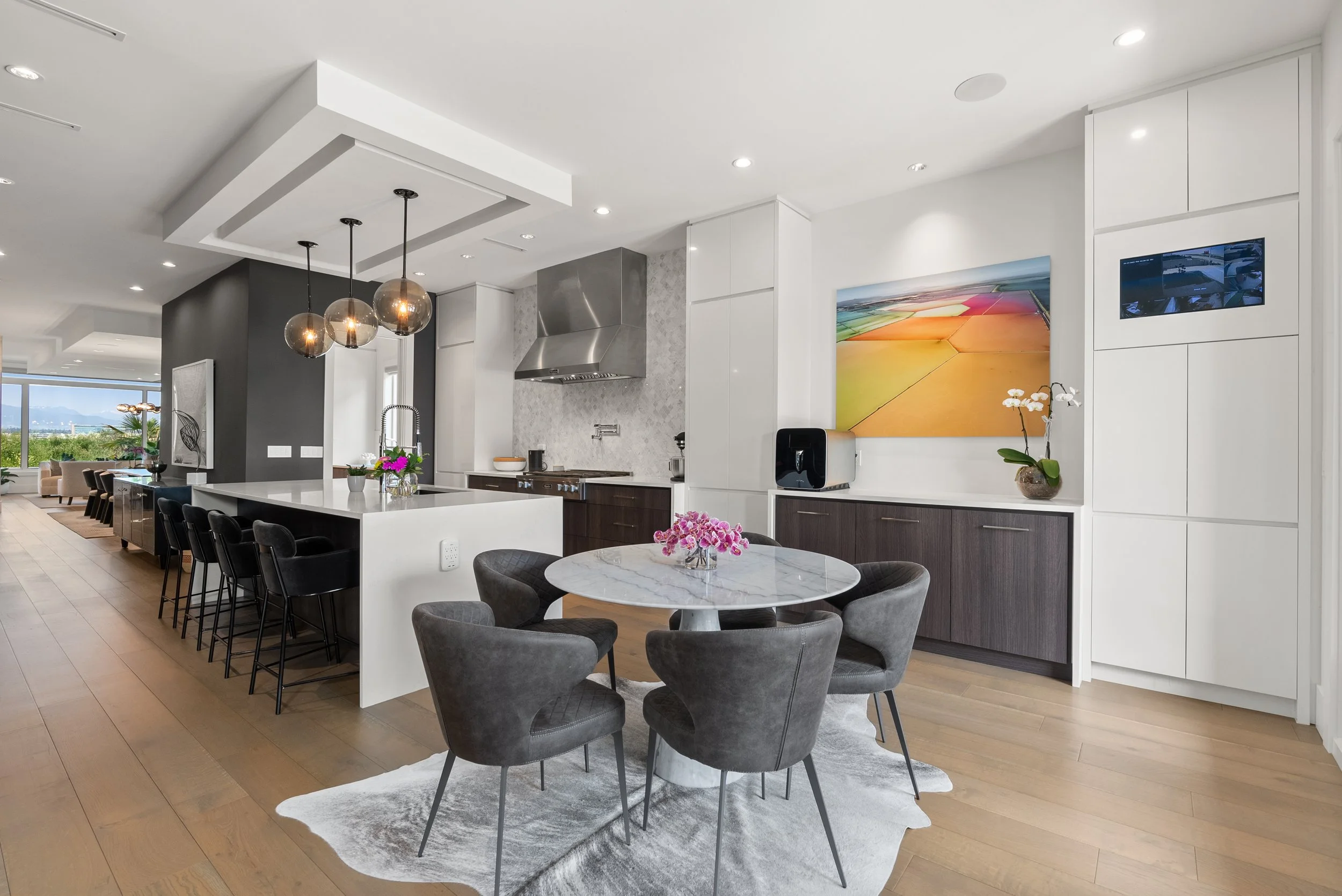 Modern open-concept kitchen and dining area with white cabinets, marble countertops, black chairs, and hanging pendant lights, with a living room and large window view in the background.
