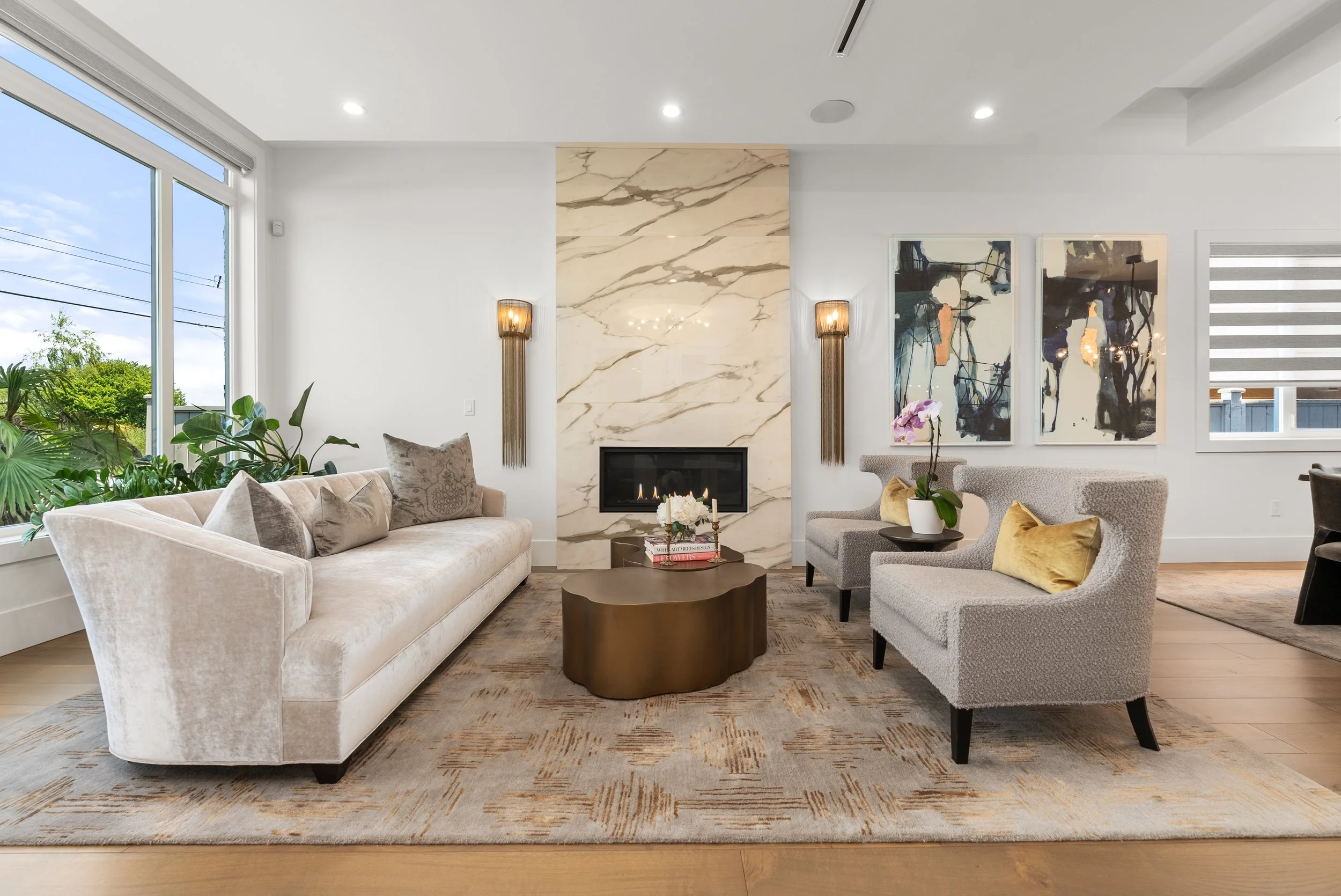 Modern living room with large window, beige sofa, two gray armchairs with yellow pillows, a marble fireplace, wall art, and a coffee table with flowers.