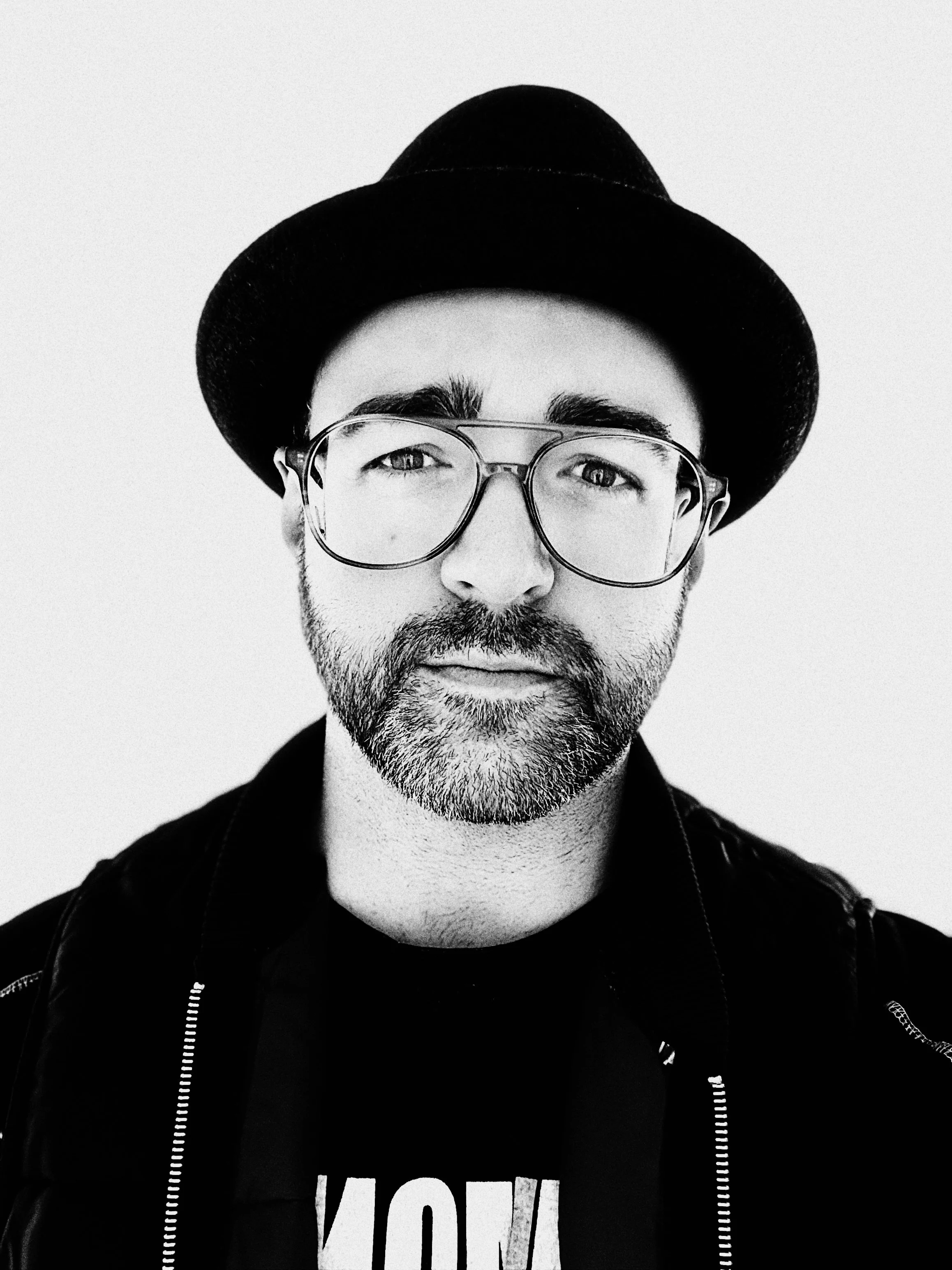 A man with glasses, a beard, and a mustache wearing a black hat and a black jacket, smiling slightly, against a plain light background.