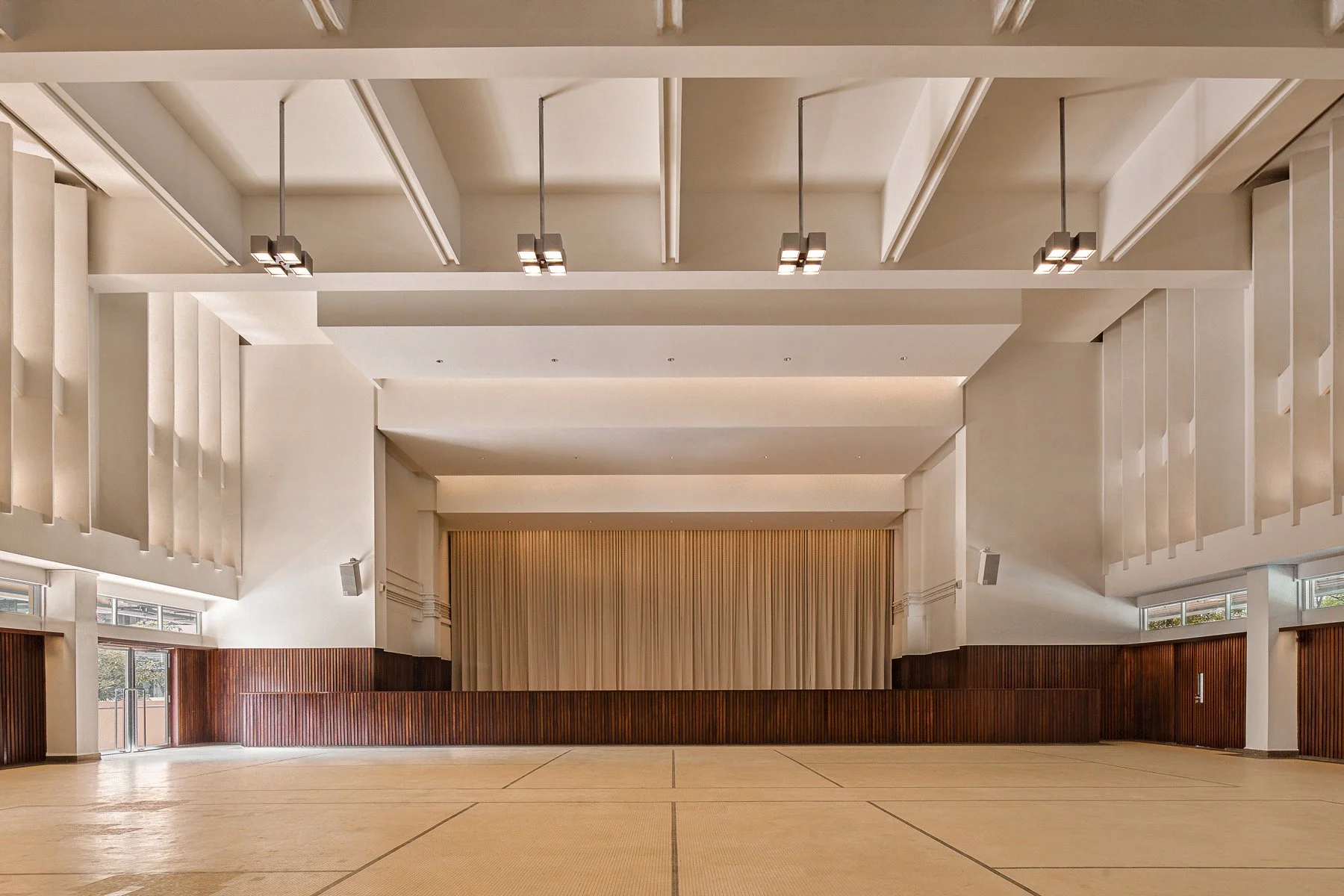 Empty indoor auditorium with wooden stage, beige walls, curtains, and overhead lights.