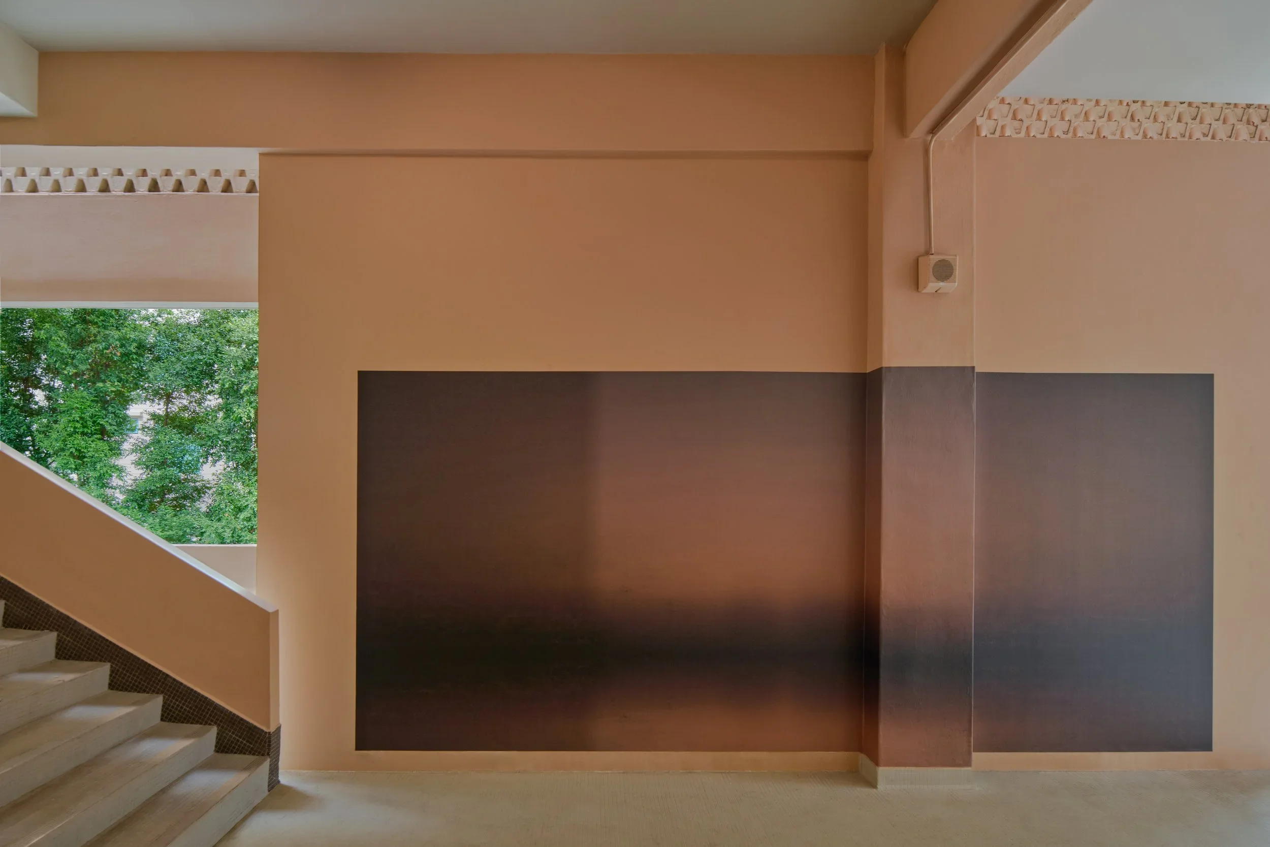 Interior view of a staircase with a beige and dark brown wall design, with a window showing green trees outside, and a small speaker mounted on the wall.