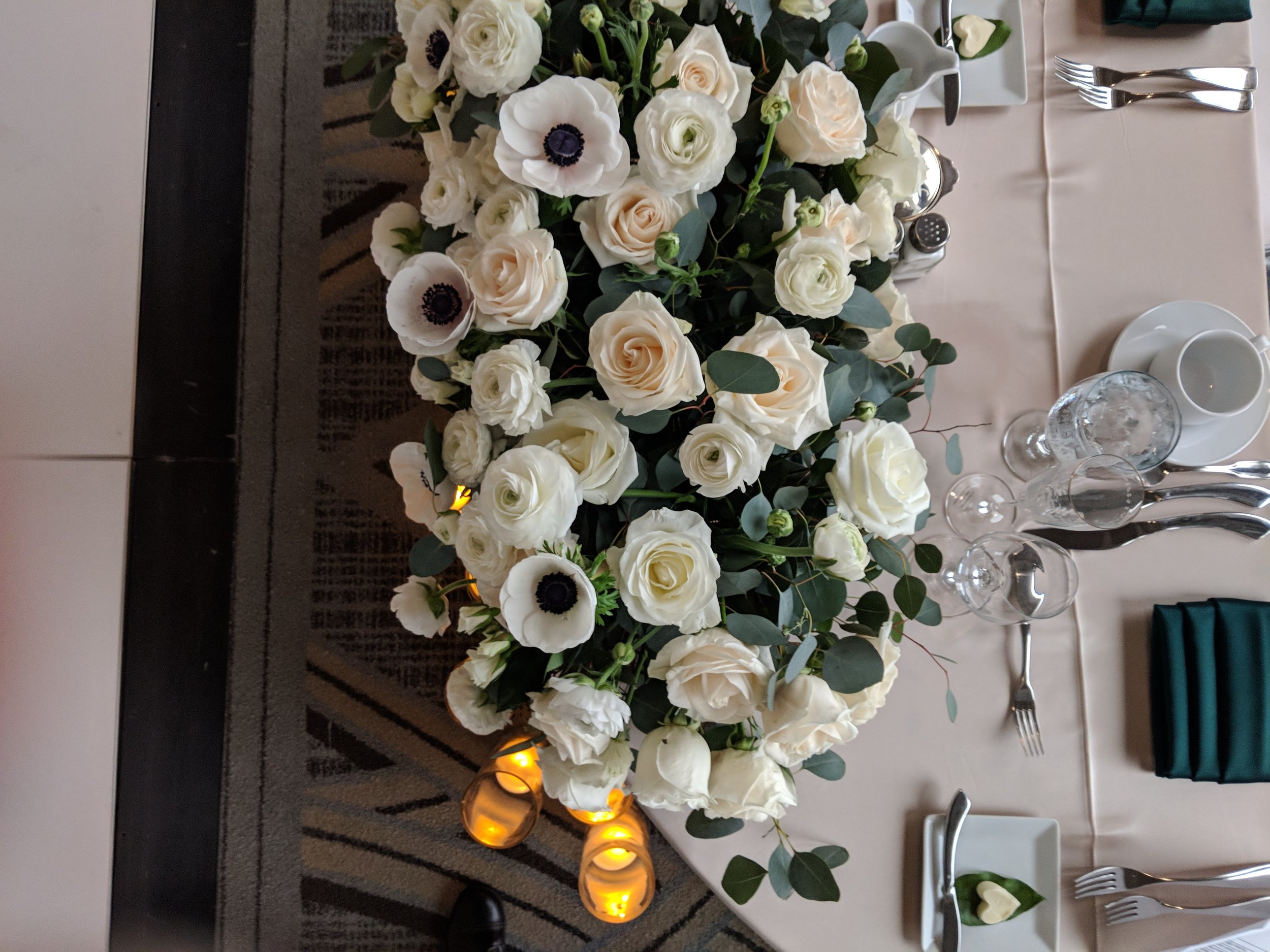 Elegant table setting with a large floral centerpiece featuring white and cream roses and anemones, accented with greenery. The table includes several place settings with cutlery, glasses, and napkins, along with glowing candles underneath.