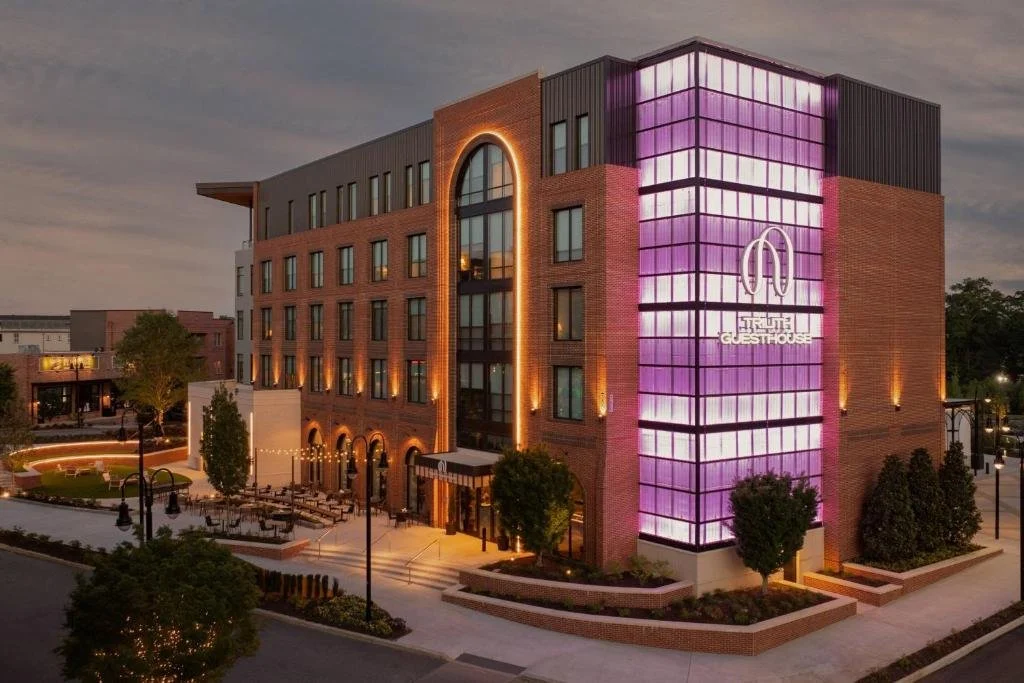 Modern four-story brick hotel building with purple neon lighting, trees, outdoor seating, and street lamps at dusk.-venues near me-local venues-interior scapes events by julie jenkins