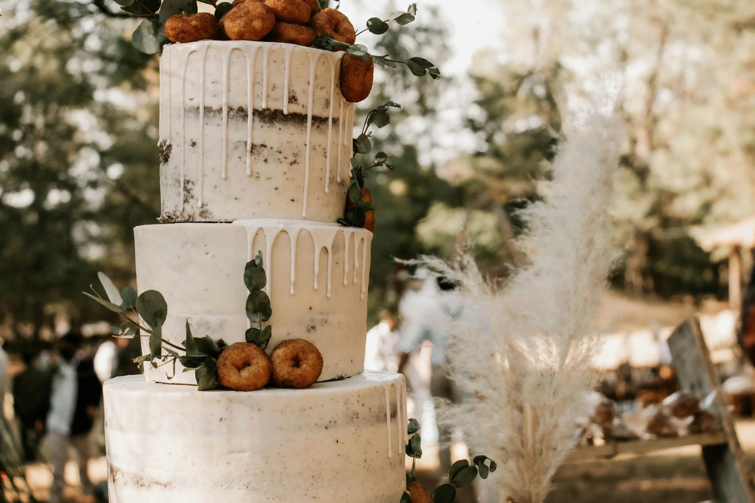 Three-tiered wedding cake decorated with greenery and donuts, with white drip icing, outdoors with trees in the background.  Seasonal decor installation and event planning