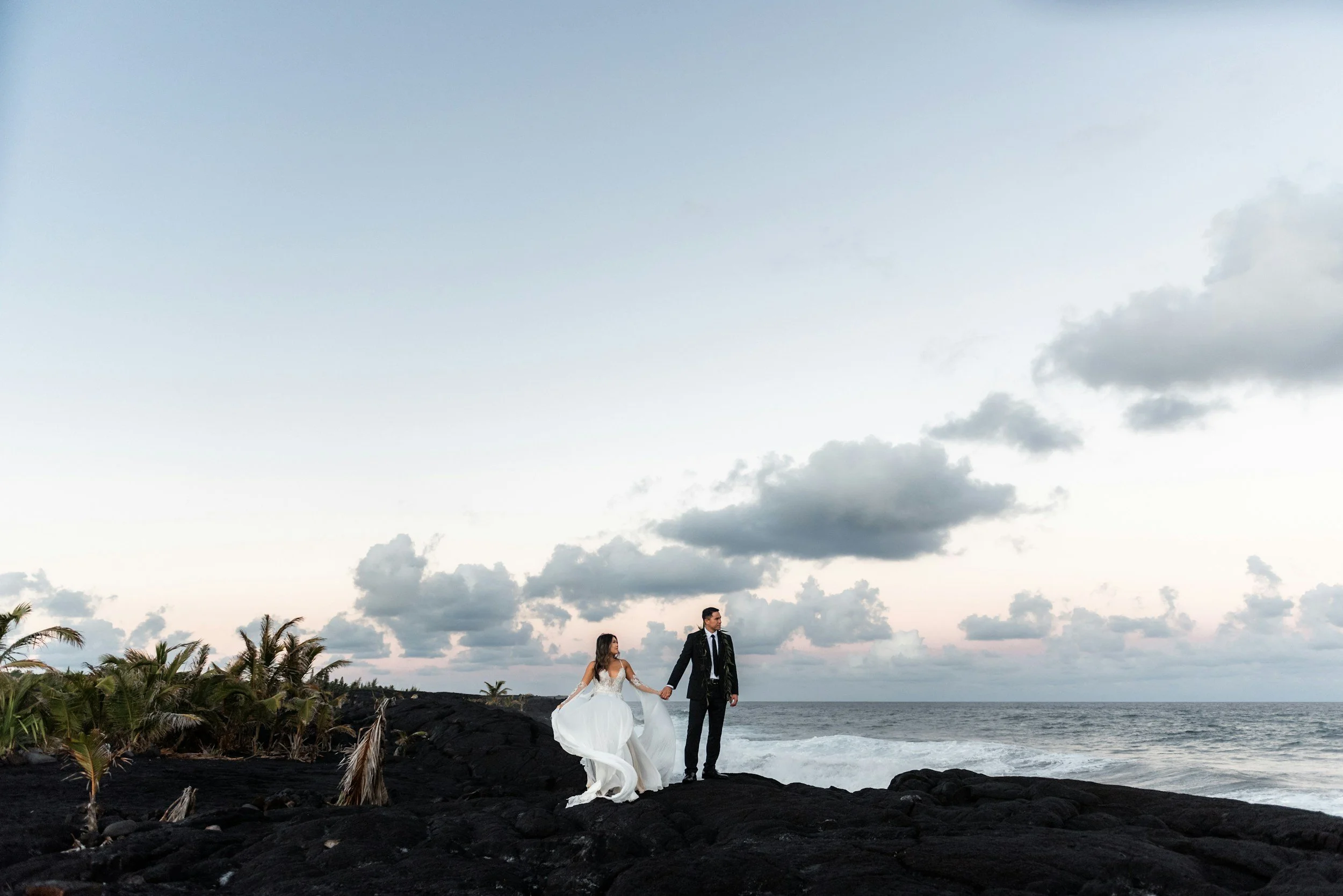 A bride and groom walking hand in hand along a rocky beach with palm trees in the background, under a partly cloudy sky.  promoting interior scapes and events by Julie Jenkins