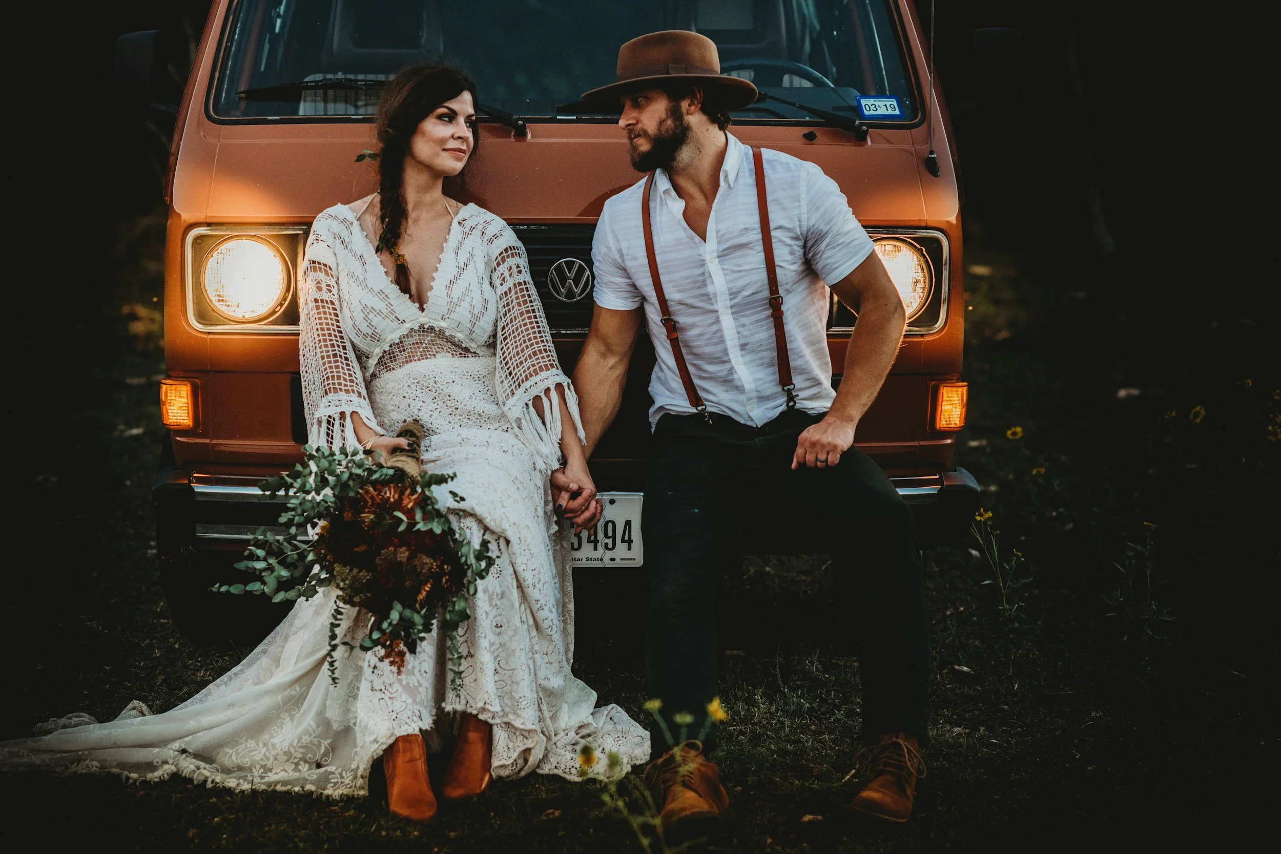 A couple dressed in vintage clothing sitting in front of an orange Volkswagen van, holding hands. The woman is wearing a long white lace dress with crochet details and orange boots, holding a bouquet of flowers. The man is wearing a white striped shirt, dark pants, suspenders, and a wide-brimmed hat. They are outdoors in a natural setting at sunset.