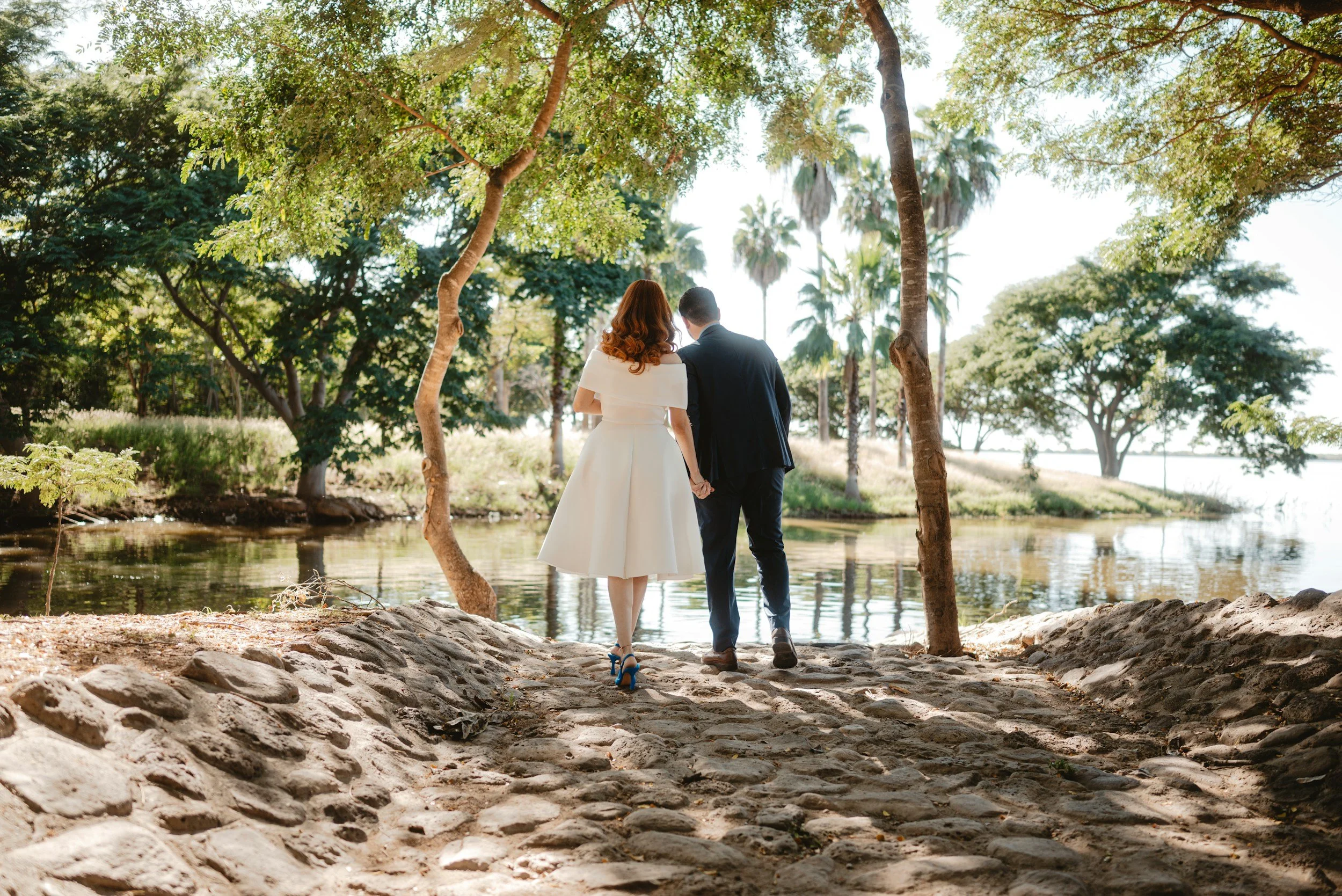 A couple dressed in wedding attire walking hand in hand by a lake in a wooded park during daytime.