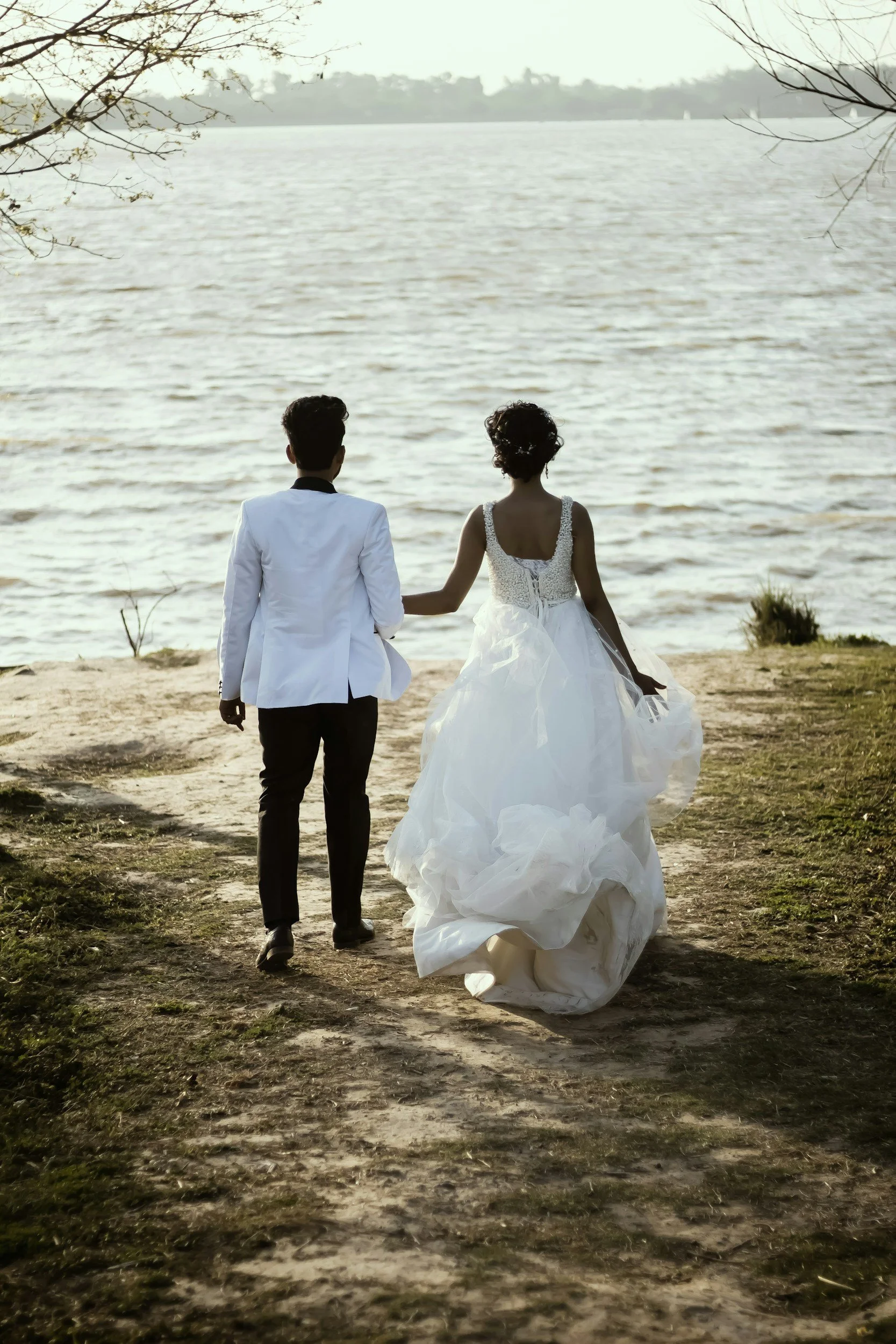 A bride and groom walking hand in hand along a lakeside path, with the bride in a white wedding dress and the groom in a white suit jacket, surrounded by trees, near a body of water.
