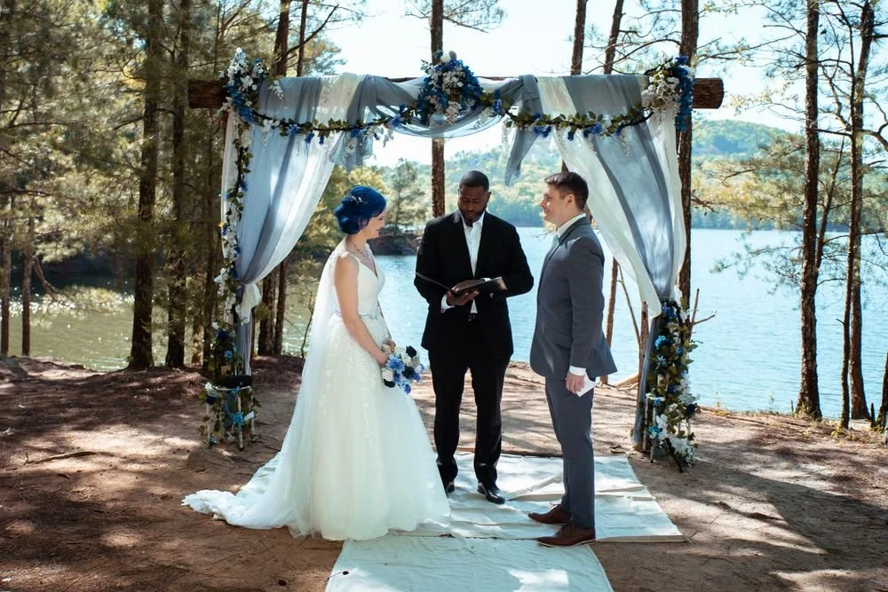A couple gets married outdoors by a lake under a decorated wooden arch with draped fabric and flowers, officiated by a person in a black suit.  Red top mountain state park GA-Interior Scapes, Events by Julie Jenkins event and wedding planning