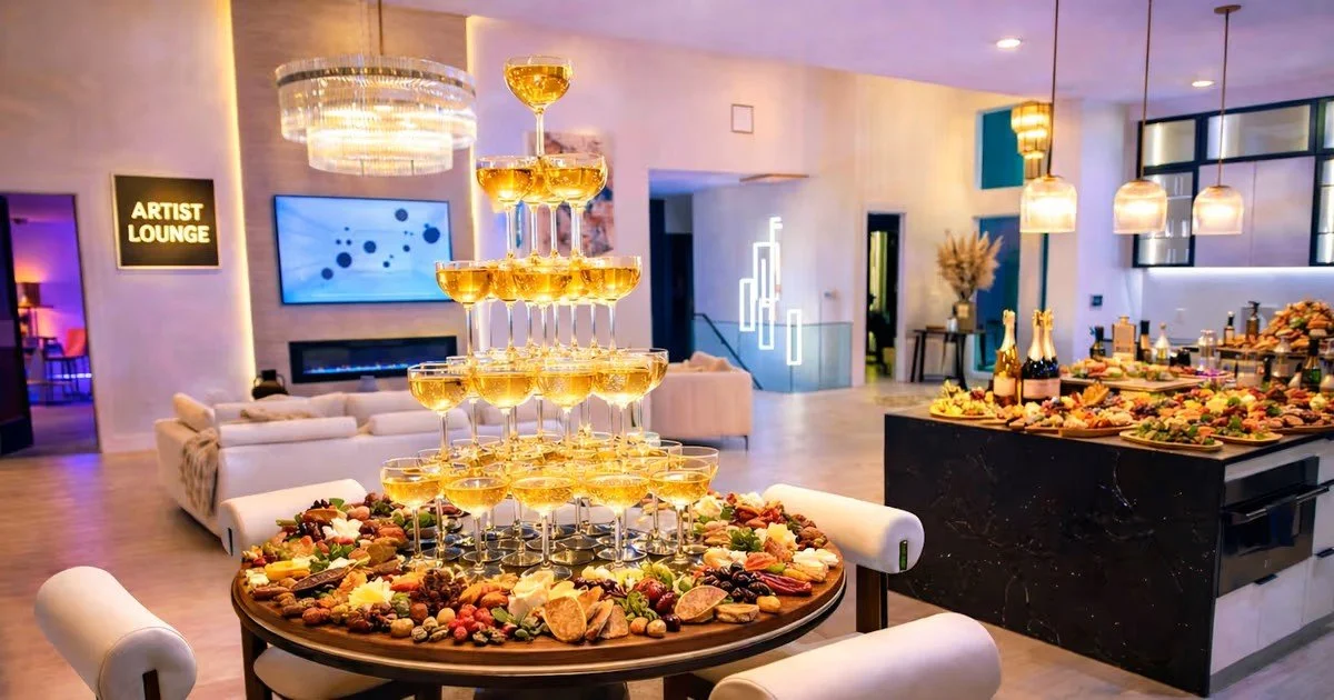 Elegant party room with a champagne tower, cheese and charcuterie spread, and modern decor including a sign that says 'Artist Lounge'.