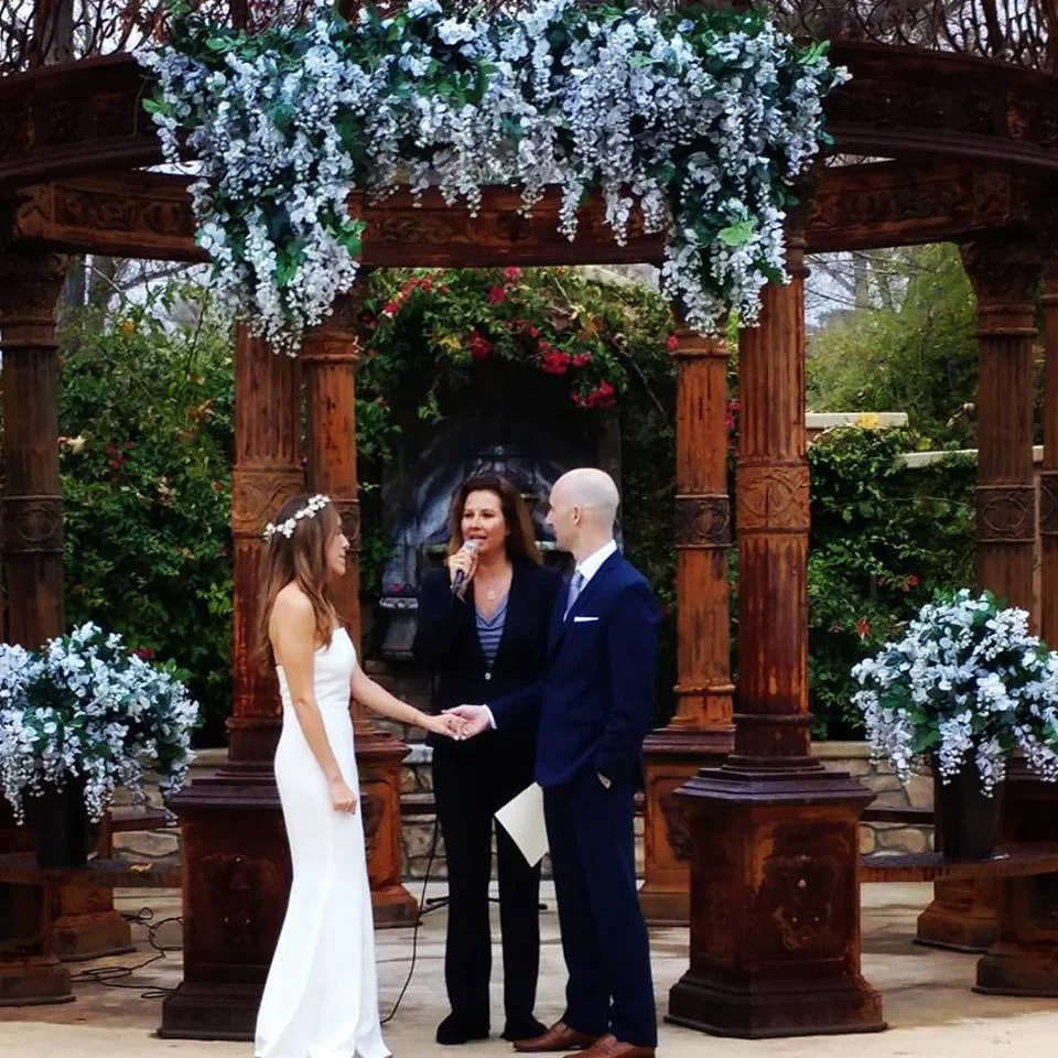 A wedding ceremony outdoors with a woman officiant, a bride in a white dress, and a groom in a navy suit, standing under a decorated wooden arch with white flowers, holding hands and exchanging vows.   Interior Scapes Ceremony Styling by Julie Jenkin