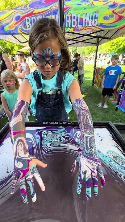 A girl with face paint and body paint on her arms is creating marbled designs with swirling colors on a tray at an outdoor event. Other children in the background are watching, and a colorful tent with the word 'BODY MARBLING' is overhead.  promoting
