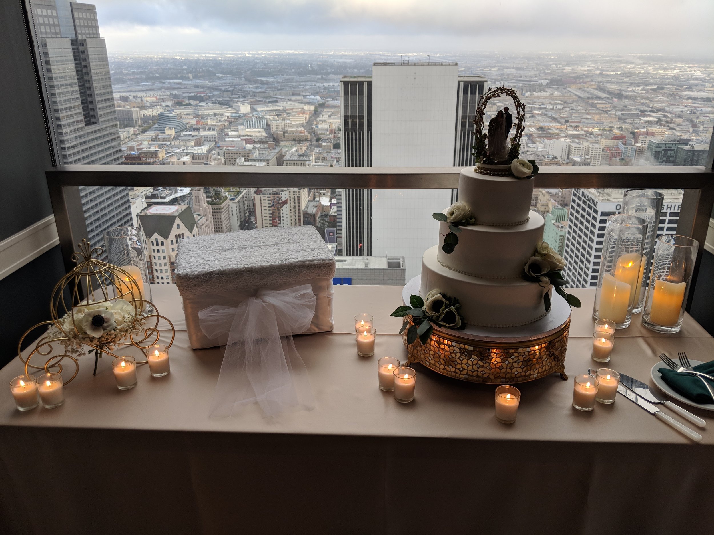 A wedding table setup in front of a city skyline. It features a three-tiered white wedding cake decorated with flowers, flanked by multiple candles. There is also a rectangular lace-covered box with a bow and a decorative wire basket with flowers. Ta