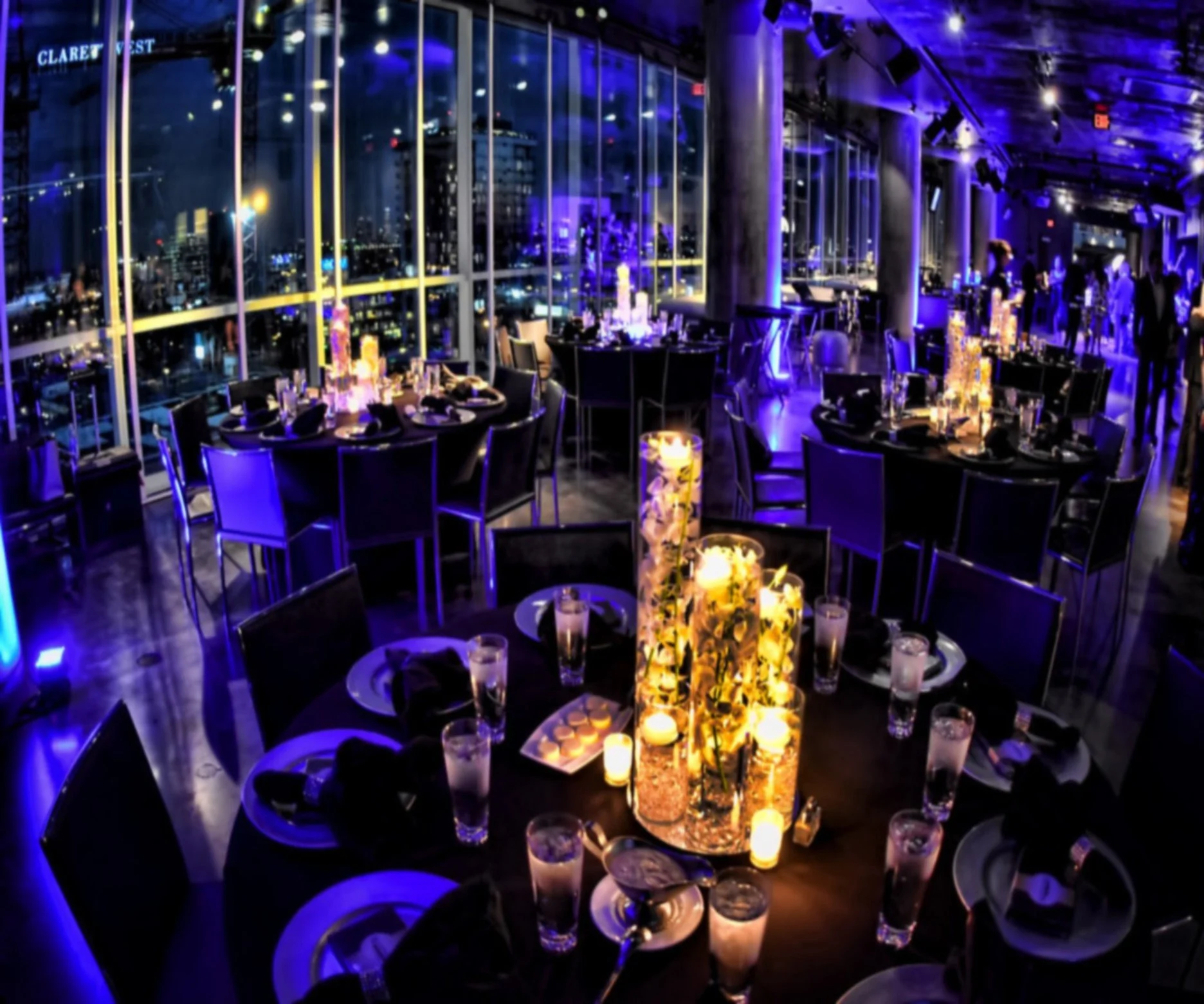 A nighttime scene inside a high-rise restaurant with large windows providing a city view. The restaurant is decorated with purple and blue lighting, illuminated candles, and floral centerpieces on tables set for dining.   promoting interior scapes an