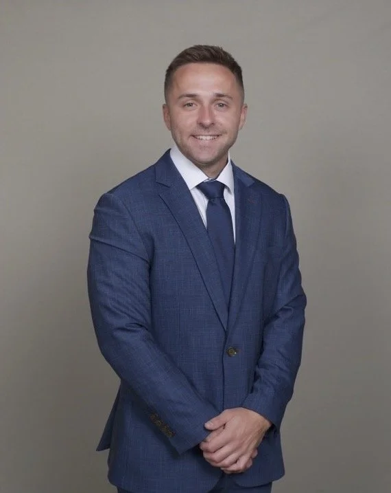 A young man in a blue suit and tie smiling against a plain gray background.