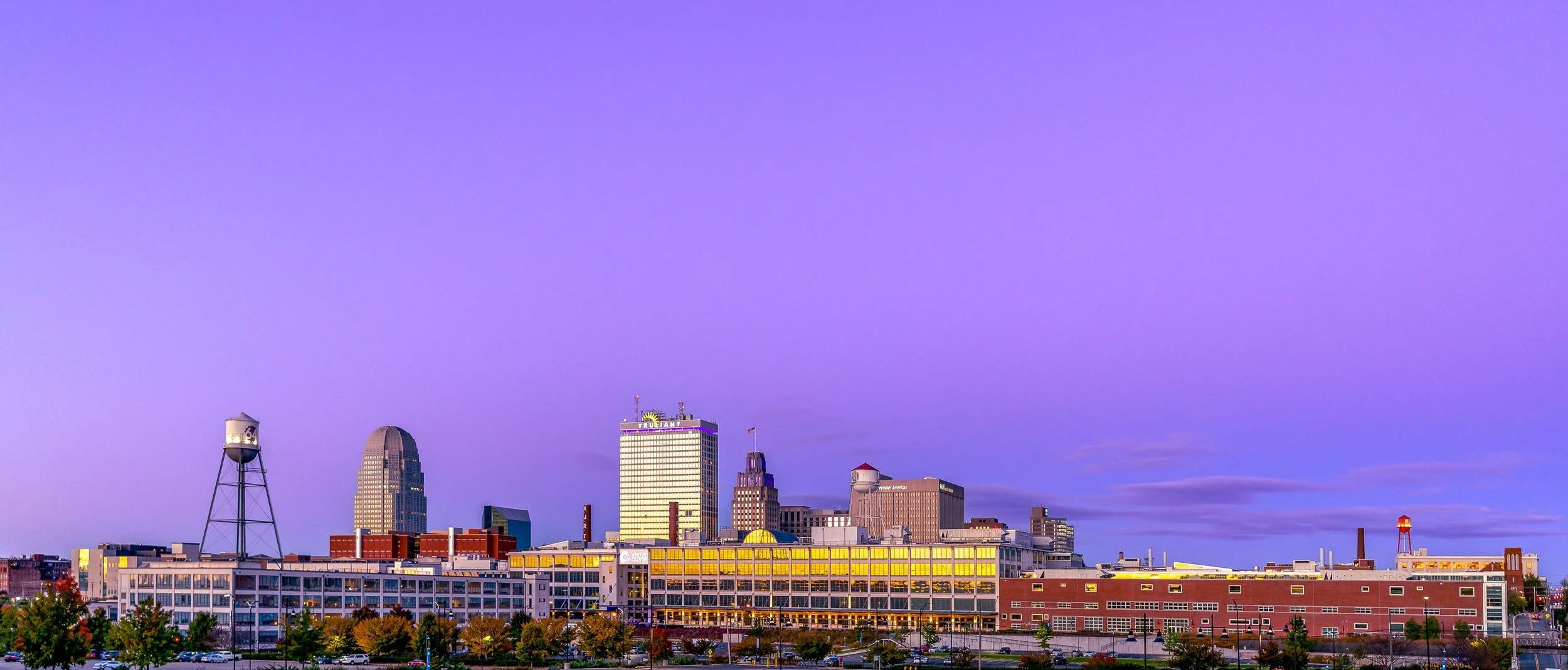 City skyline at twilight with a purple sky, featuring modern and older buildings, a prominent water tower, and a glowing branded building.