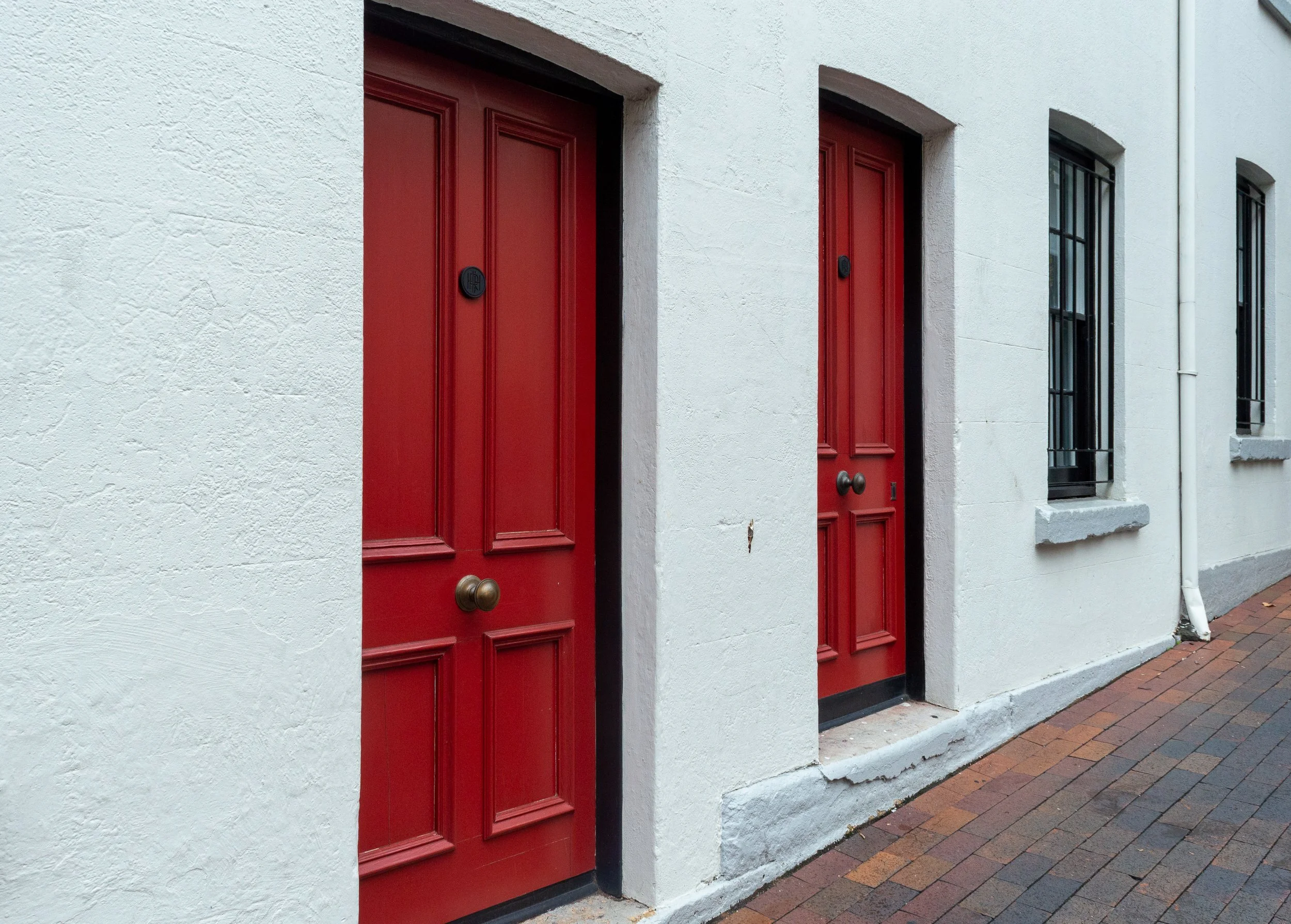 Two red doors on a white building, with adjacent windows and a brick pathway.