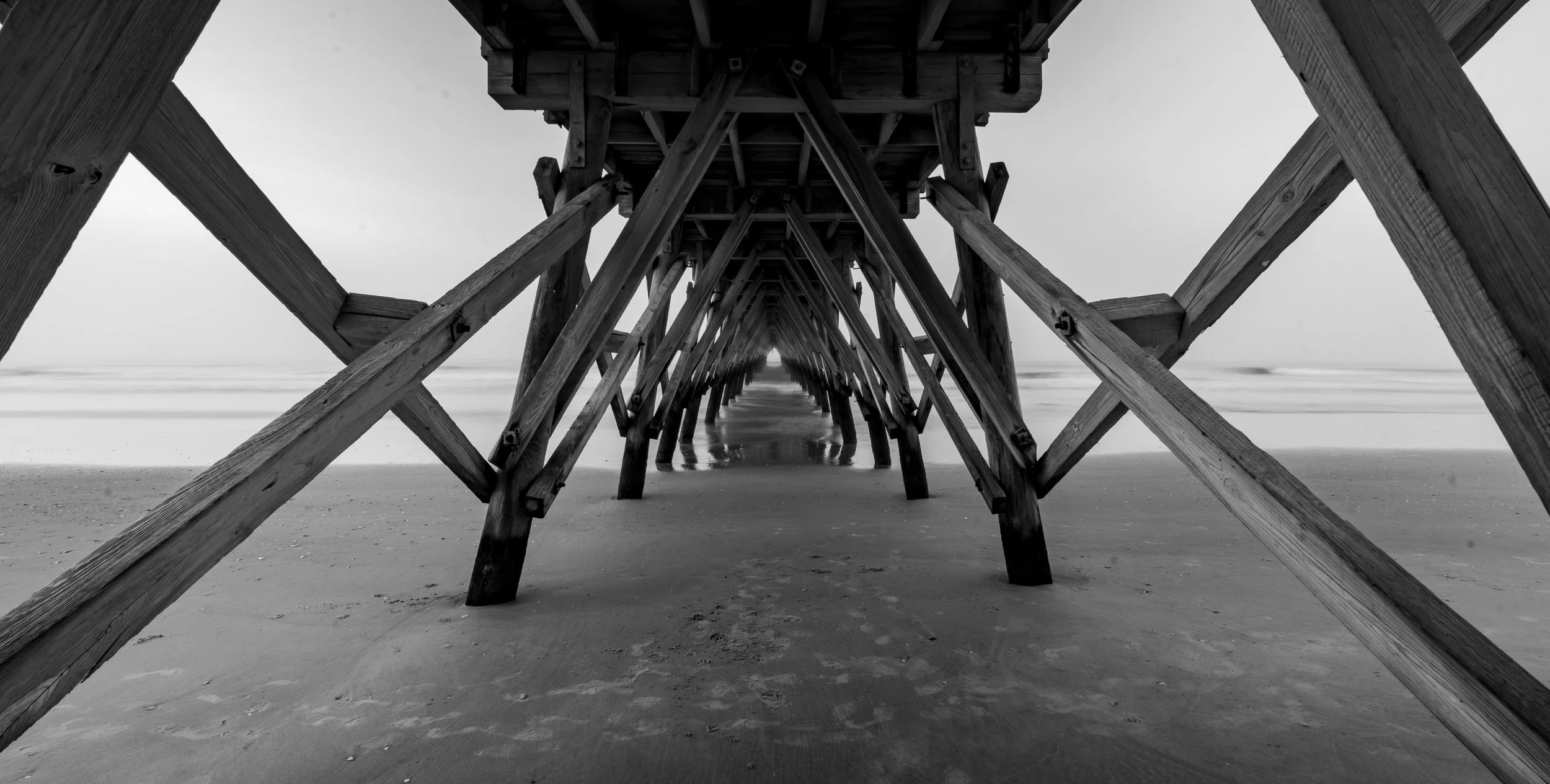 Black and white photo of a wooden pier structure on a beach, shot from underneath, showing sand and ocean in the distance.