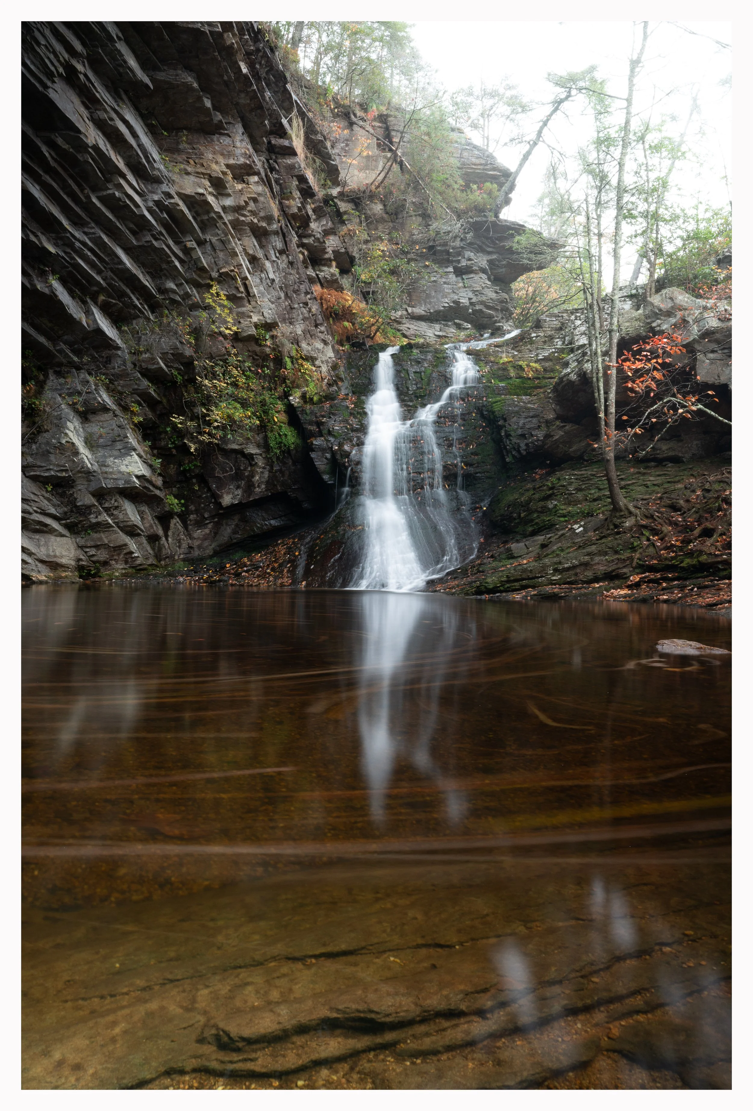A scenic waterfall cascading down a rocky cliff into a calm pool, surrounded by lush greenery and trees.